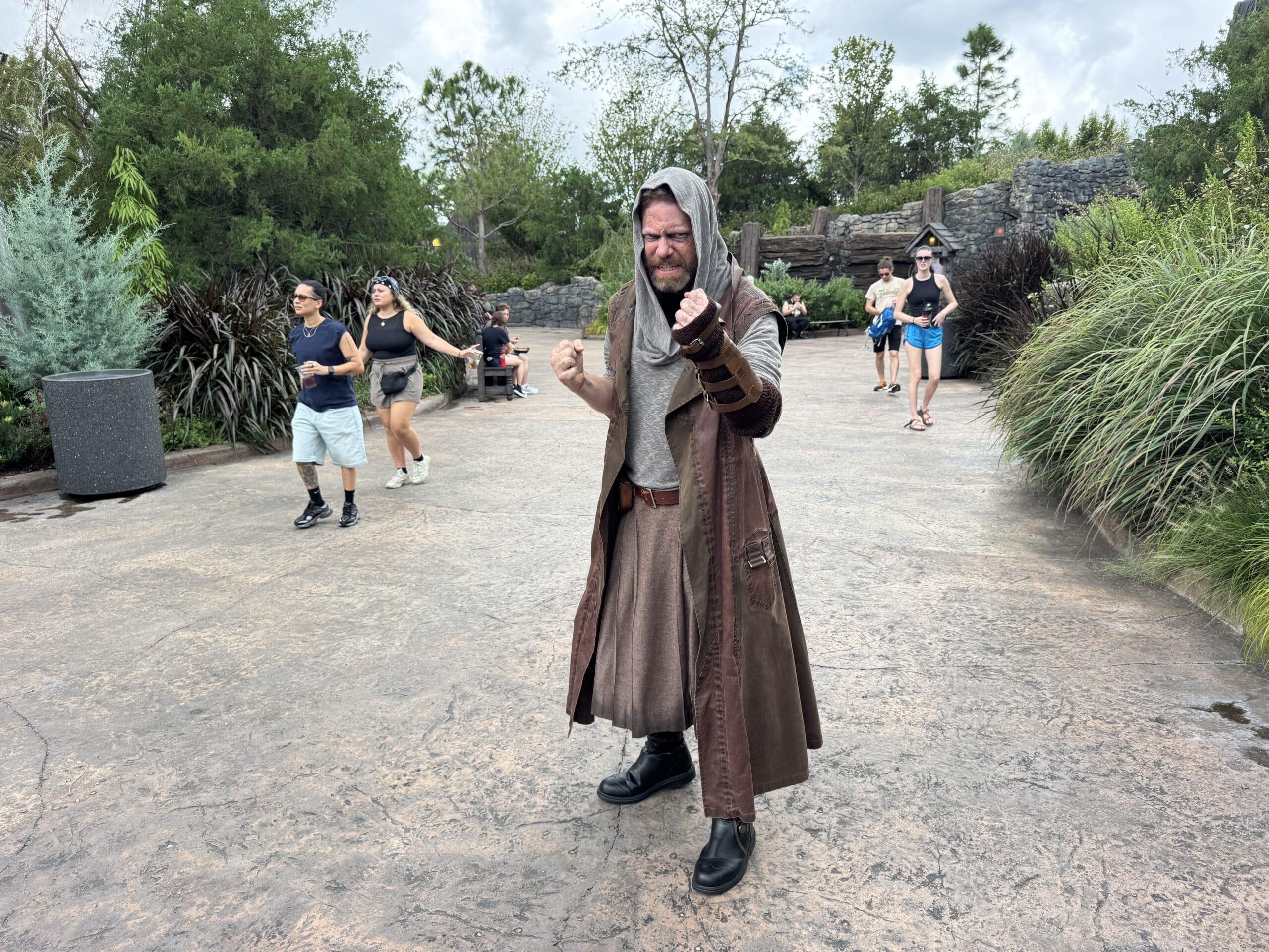 A man in a brown Jedi costume poses outdoors at Epic Universe, while people walk in the background on a theme park path.