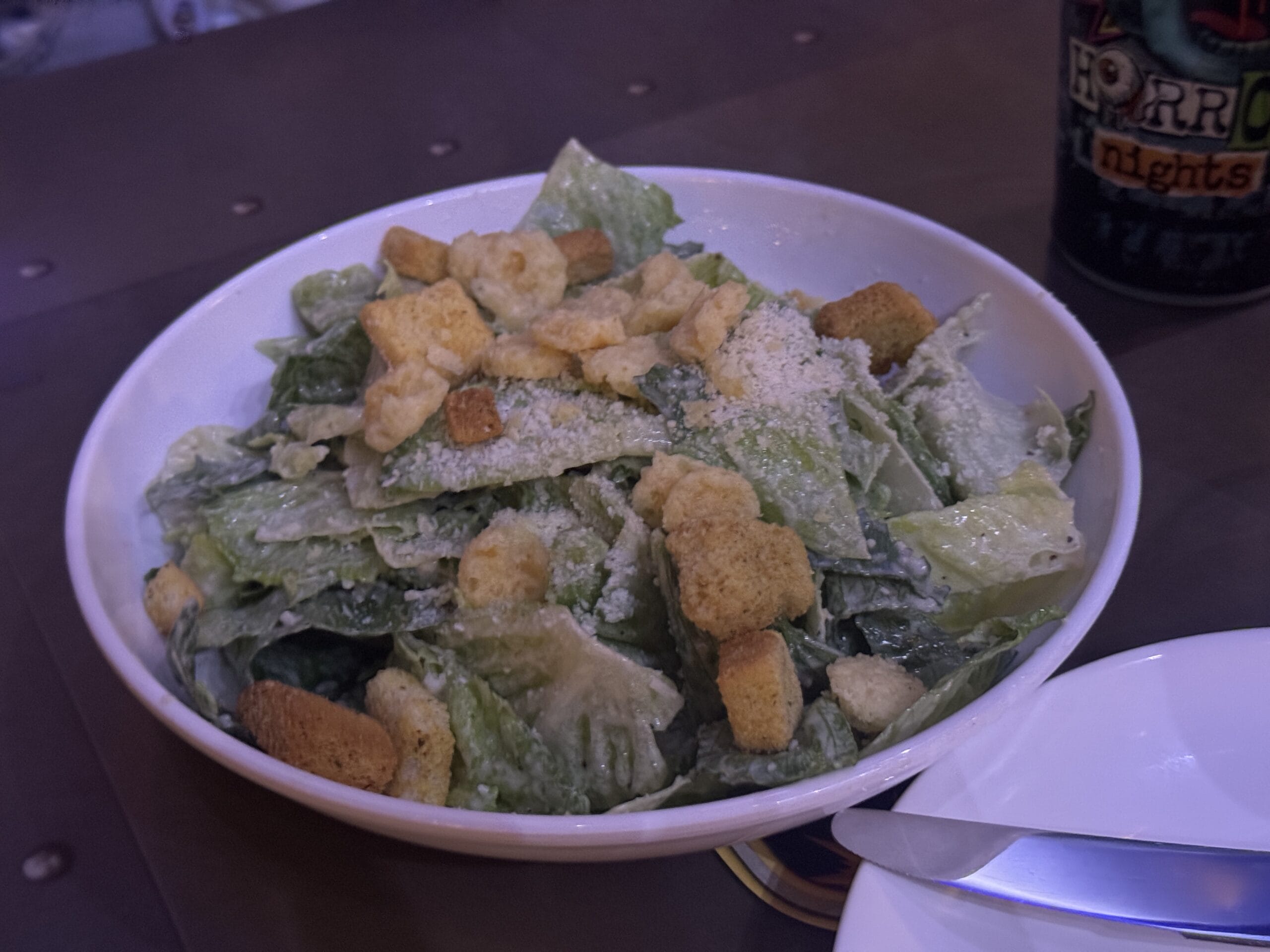 A bowl of Caesar salad with croutons and cheese, perfect comfort for a rainy day, sits on a dark table.