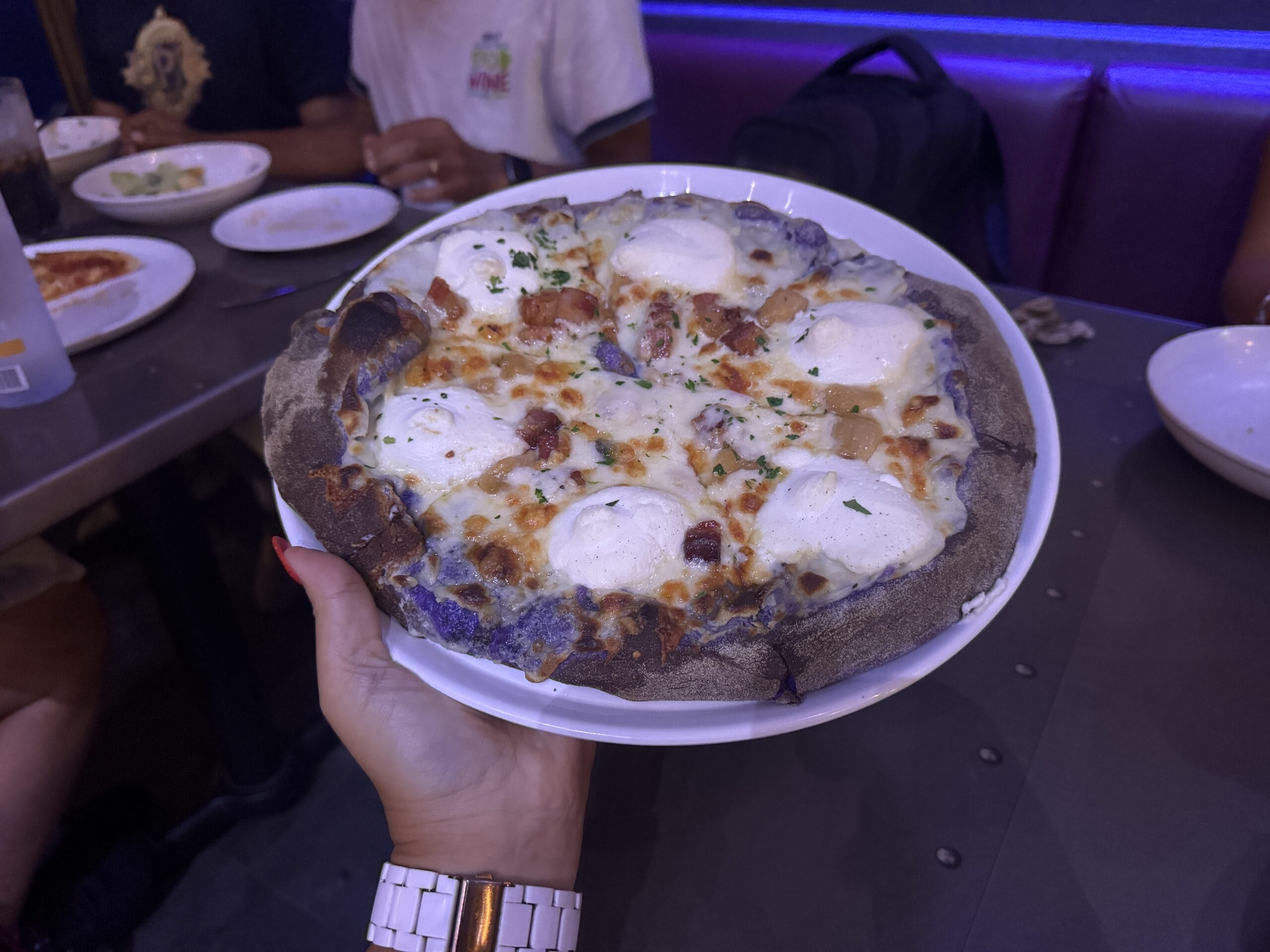 A person holds a pizza with ricotta and herbs on a dark crust, ready to enjoy at Epic Universe's restaurant setting.
