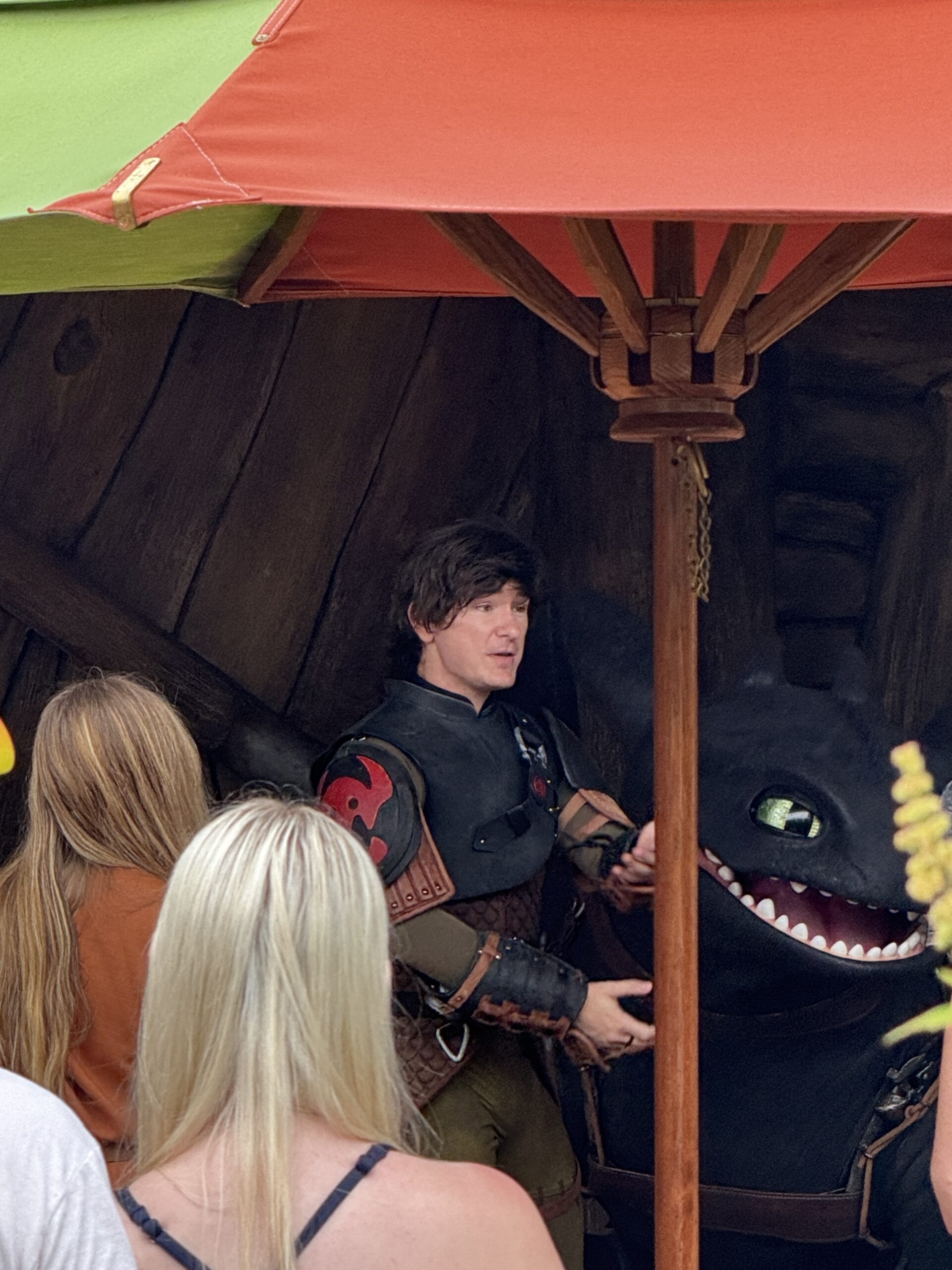 A person in medieval costume stands beside a black dragon character at Epic Universe under an orange outdoor umbrella.
