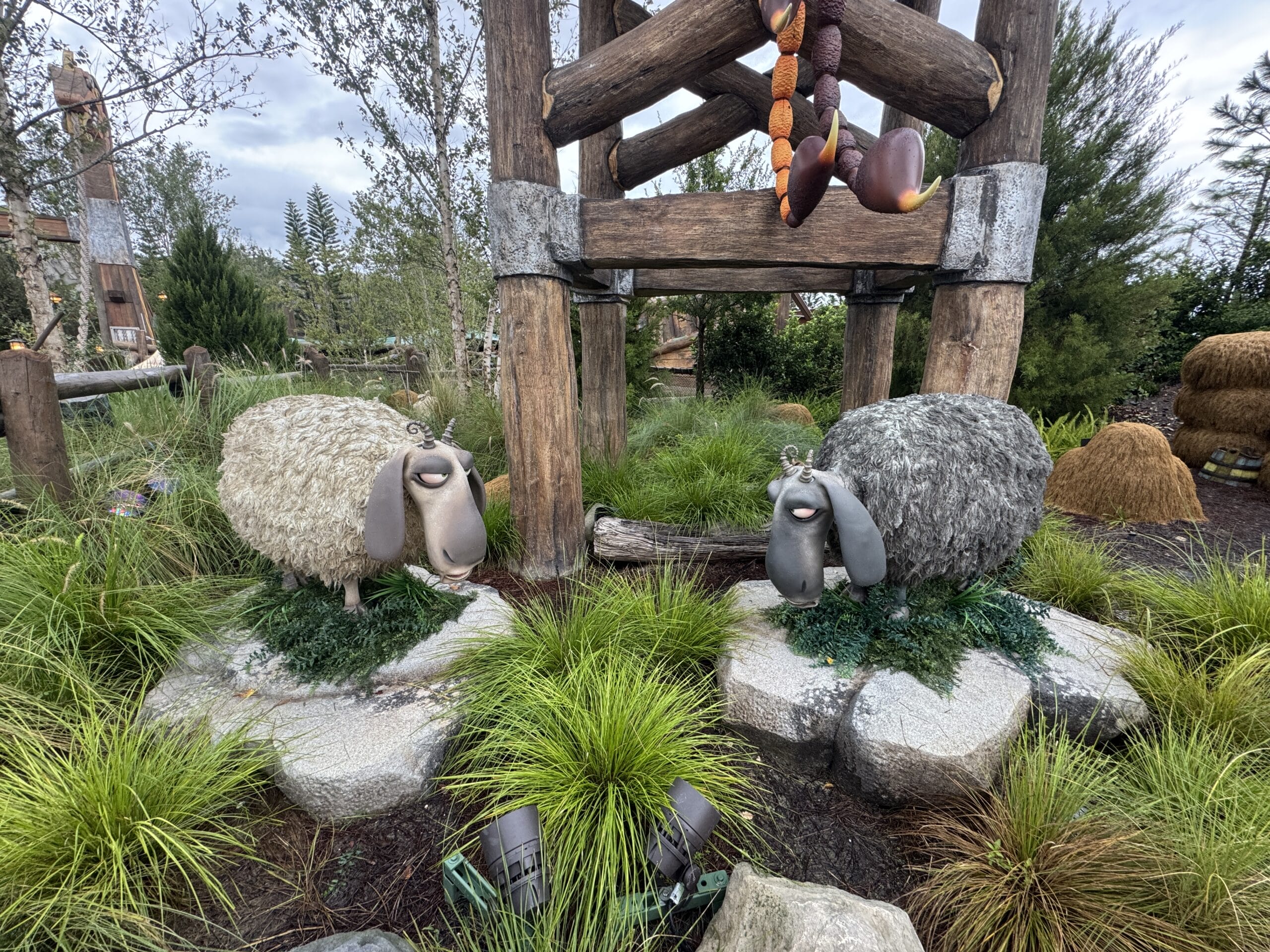 Two smiling sheep sculptures stand on stone platforms in Epic Universe’s green grass and wooden beams outdoor setting.