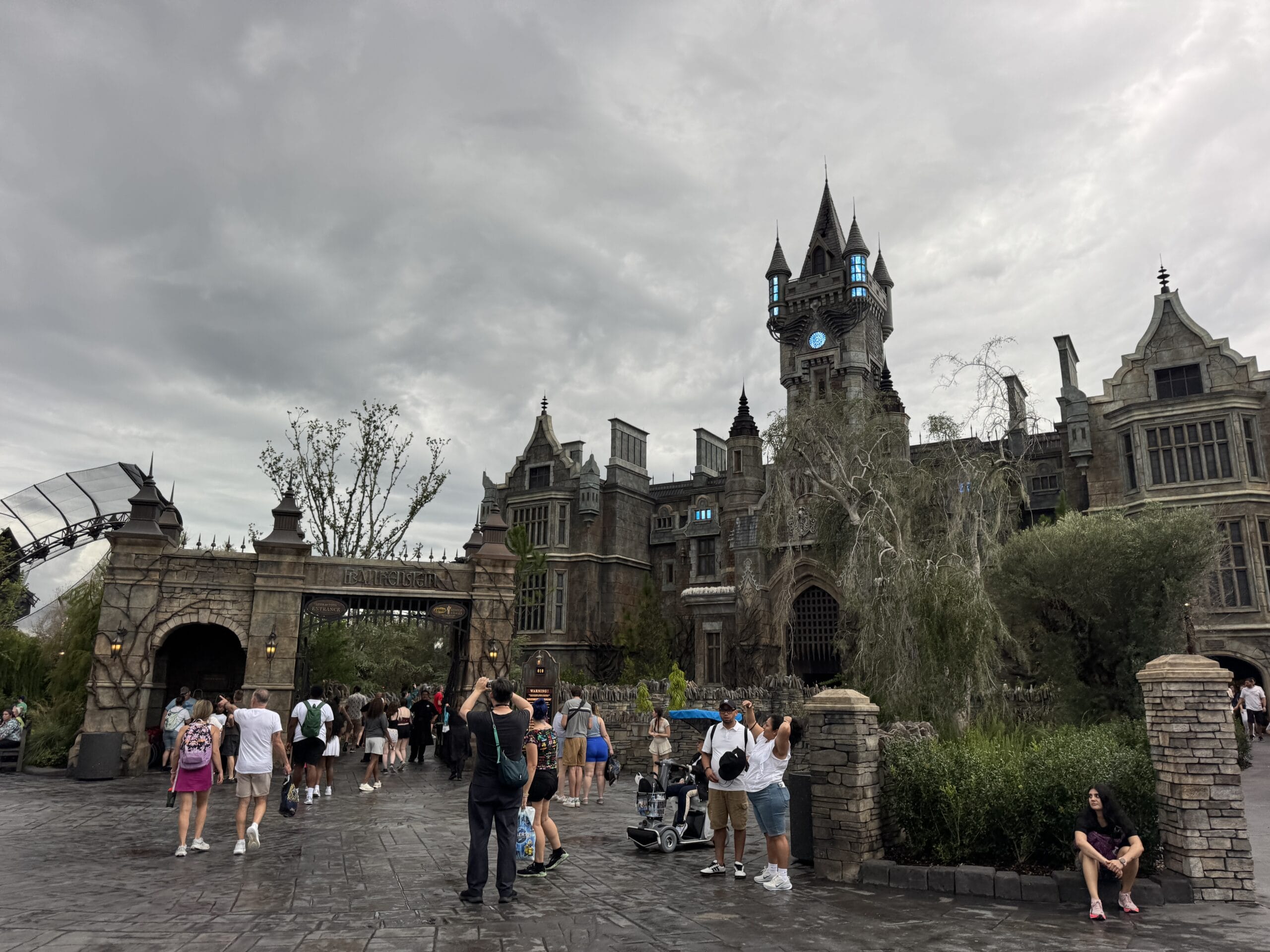 People gather outside a Gothic-style mansion with a clock tower, ready to personalize your wand on this rainy day.