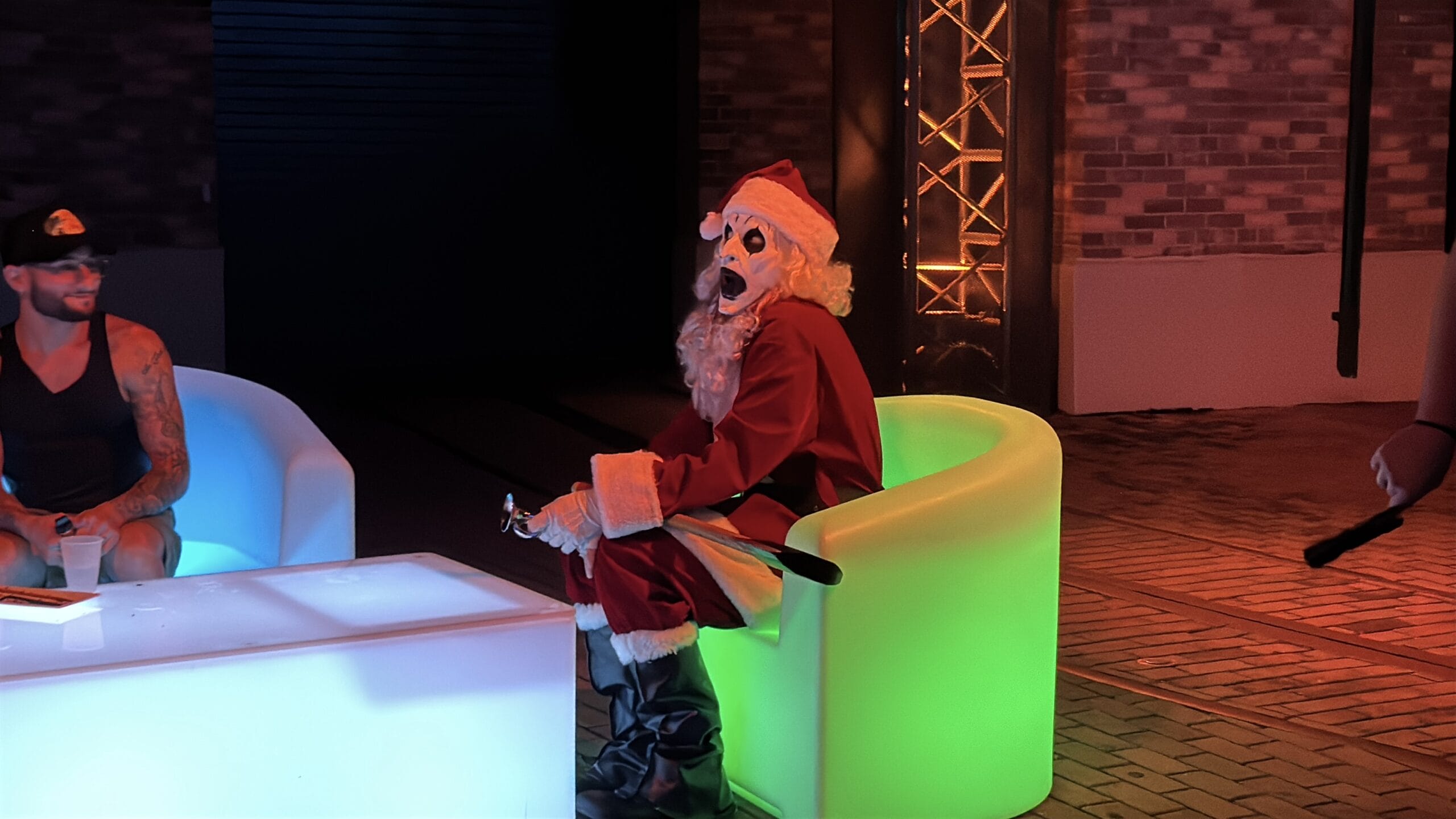 Person in a Santa suit and skull mask at Halloween Horror Night sits on a glowing green chair by a lit table at night.