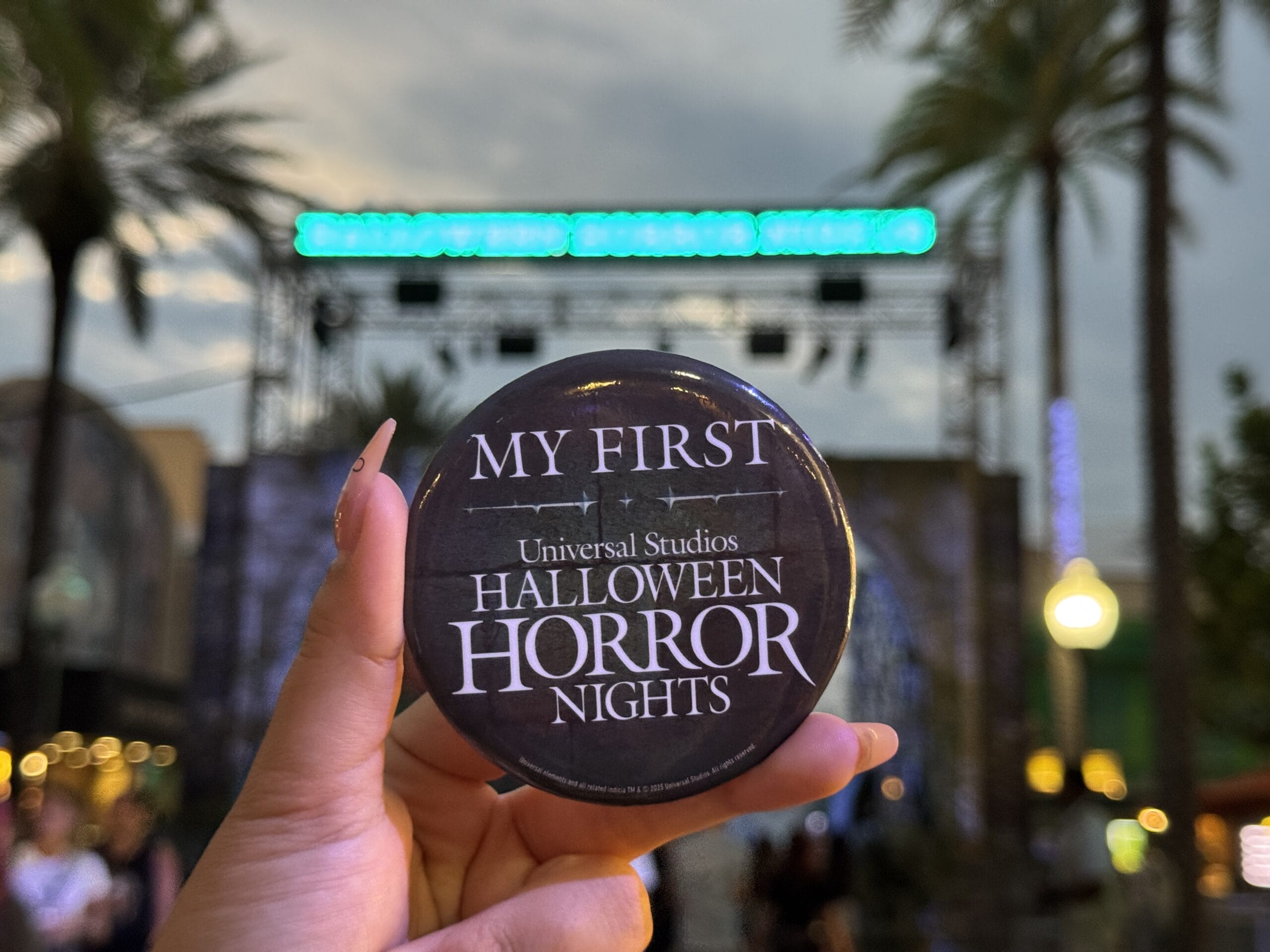 A hand holds a "My First Universal Studios Halloween Horror Night: Rush of Fear" button at an outdoor event with palm trees.