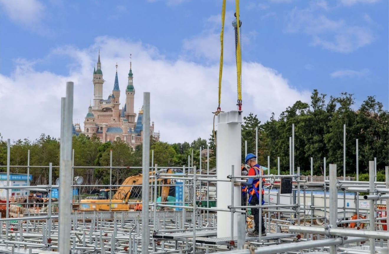 A construction worker stands among scaffolding, with Shanghai Disneyland’s themed land and a castle visible under a blue sky.