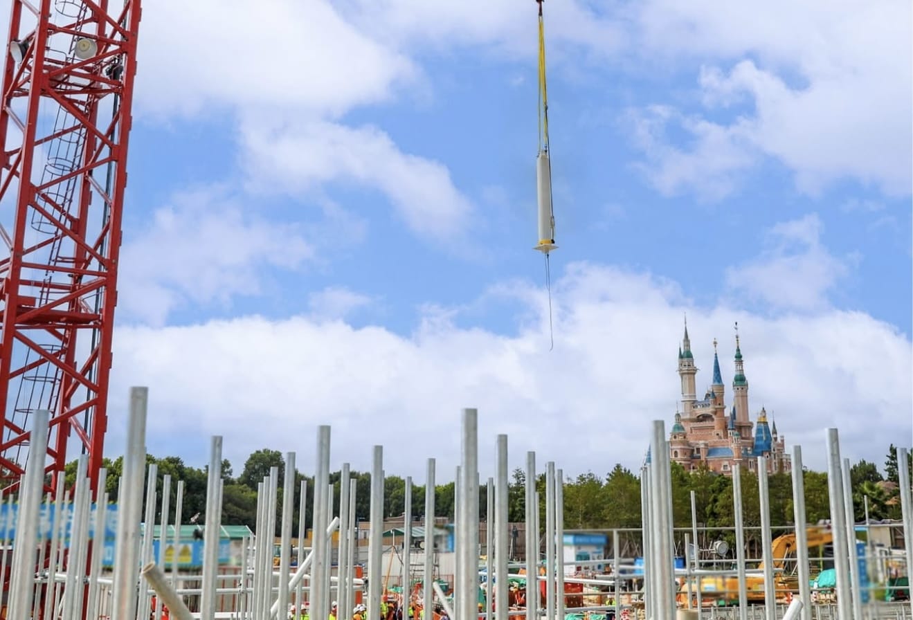 Construction site with metal poles and crane in a themed land at Shanghai Disneyland, castle under partly cloudy sky.