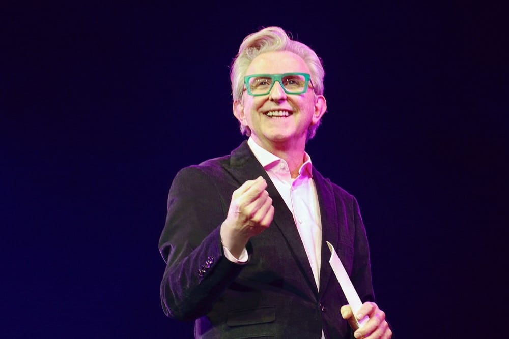 Smiling man in glasses and a suit jacket holds paper on stage under purple lights, celebrating Thomas Schumacher leaving Disney Theatrical.