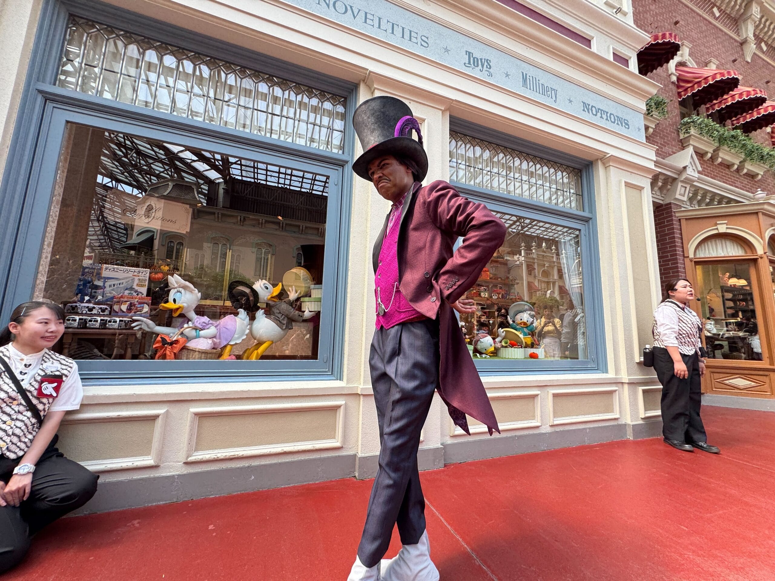 A performer in a Dr. Facilier costume and top hat strolls past a toy store window at Tokyo Disneyland, with staff nearby.