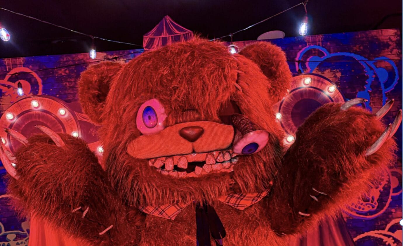 A large, creepy HamiKuma bear costume with sharp teeth and mismatched eyes prowls a Japan carnival at Universal Horror Unleashed.