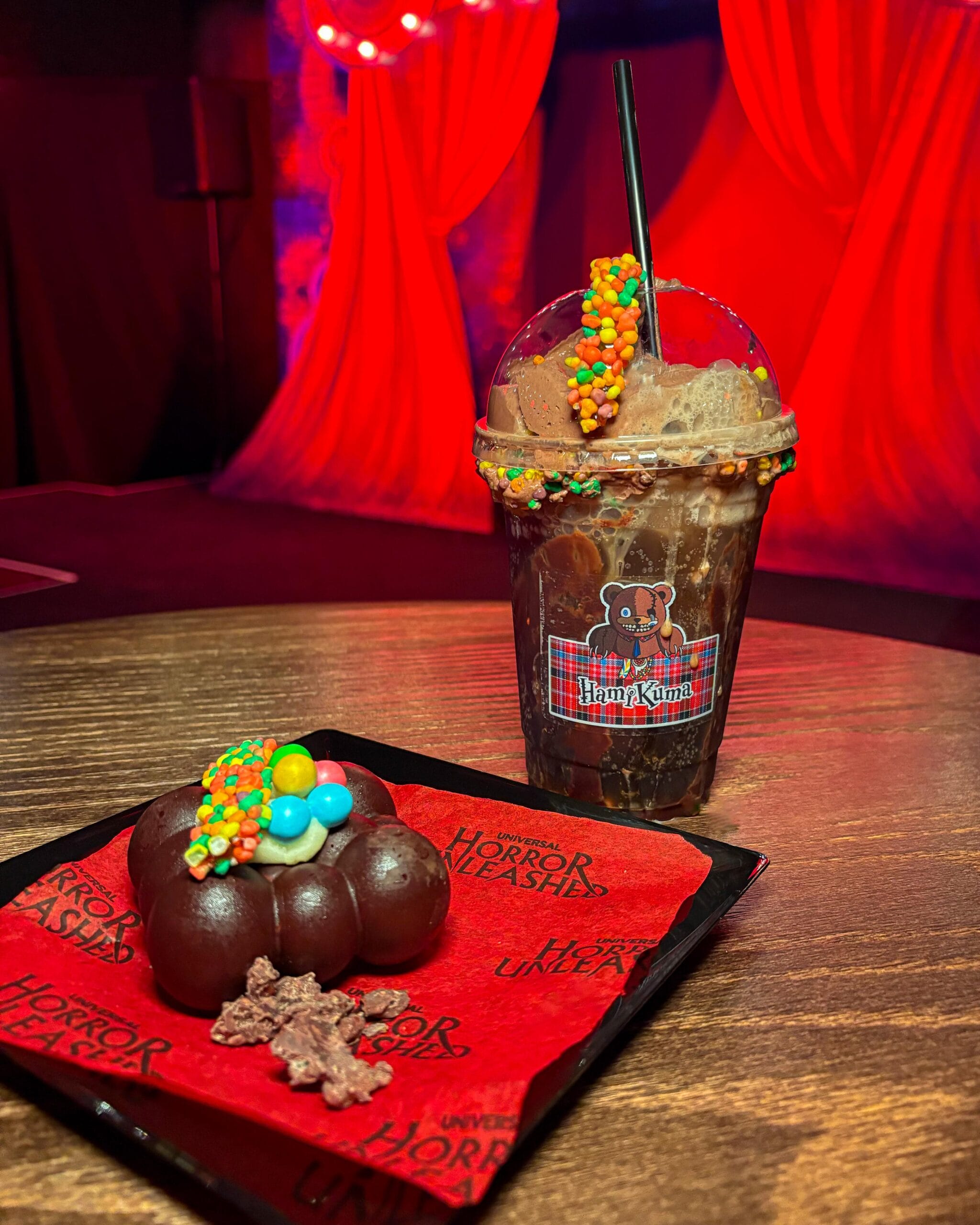 A chocolate drink and dessert with colorful candies by HamiKuma Japan, set against red curtains on a table.
