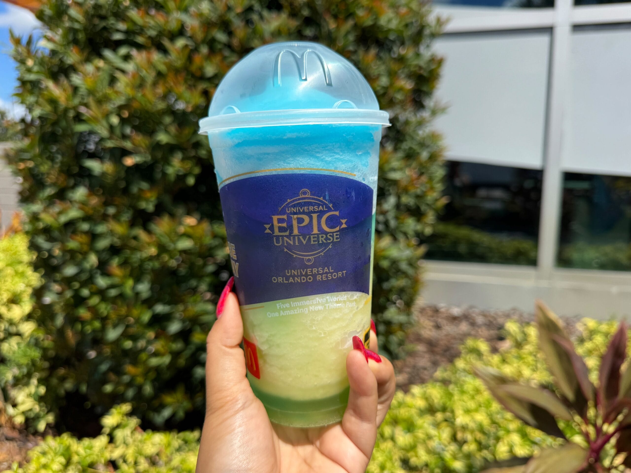 A hand holds a blue and green Epic Universe slushie cup outdoors, with foliage in the background.