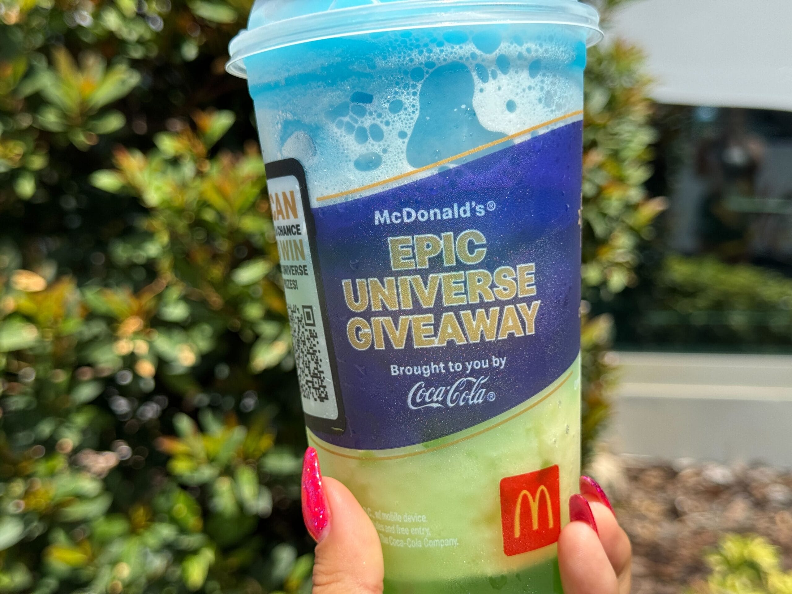 A hand holds a colorful McDonald's Slush cup promoting the Epic Universe Giveaway, with trees in the background.