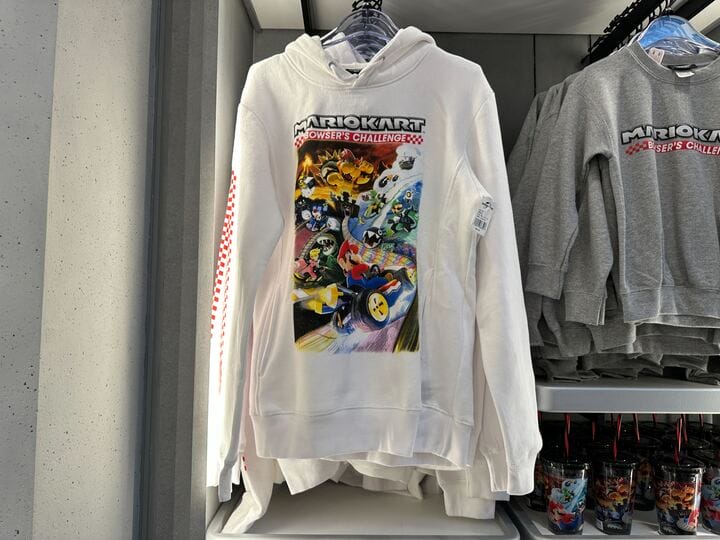 White Super Nintendo World Mario Kart: Bowser’s Challenge hoodie with colorful racers, displayed among Mario Motors merchandise.