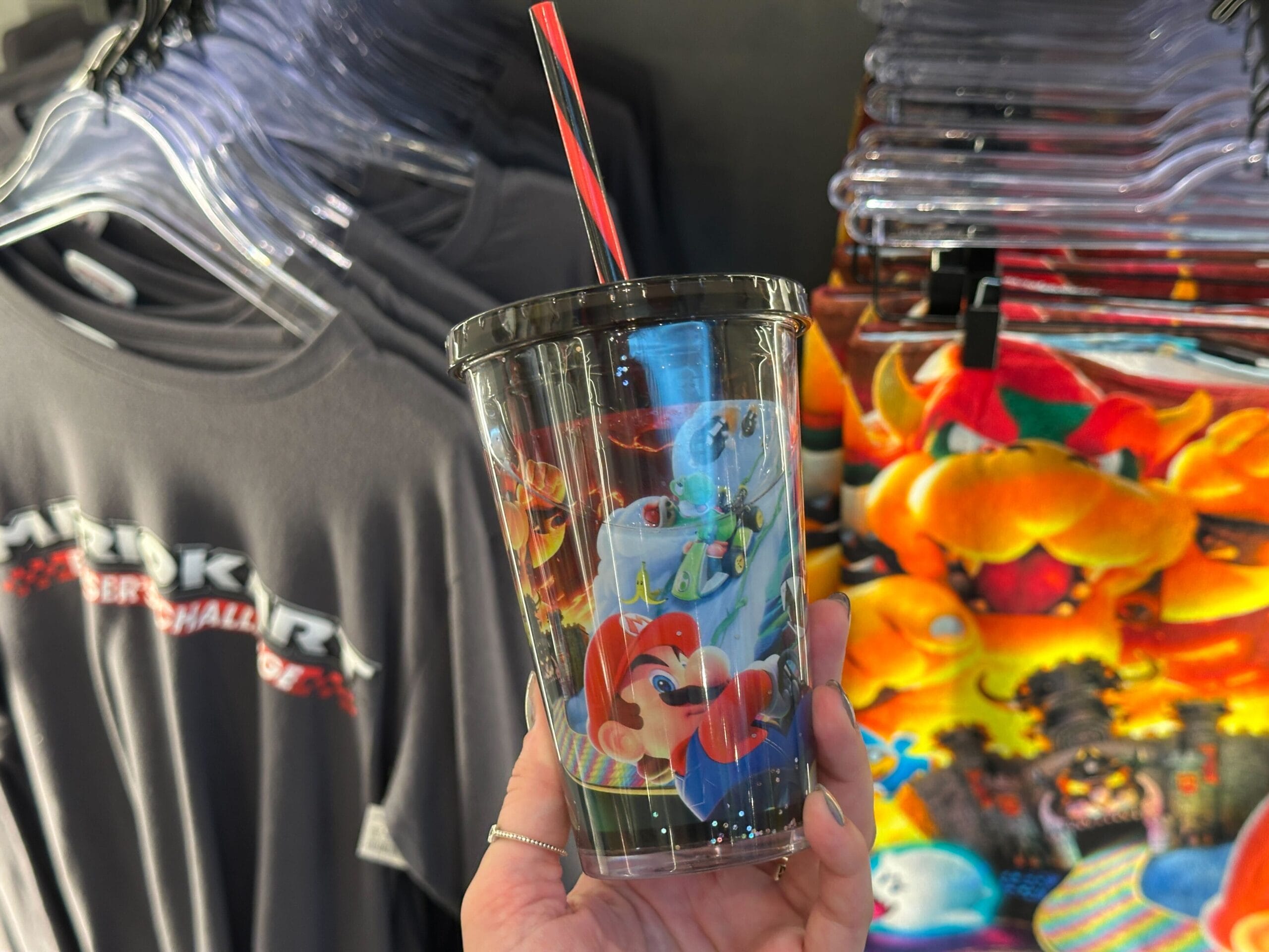 A hand holds a cup with Mario and Luigi graphics near Mario Motors merchandise at Super Nintendo World.