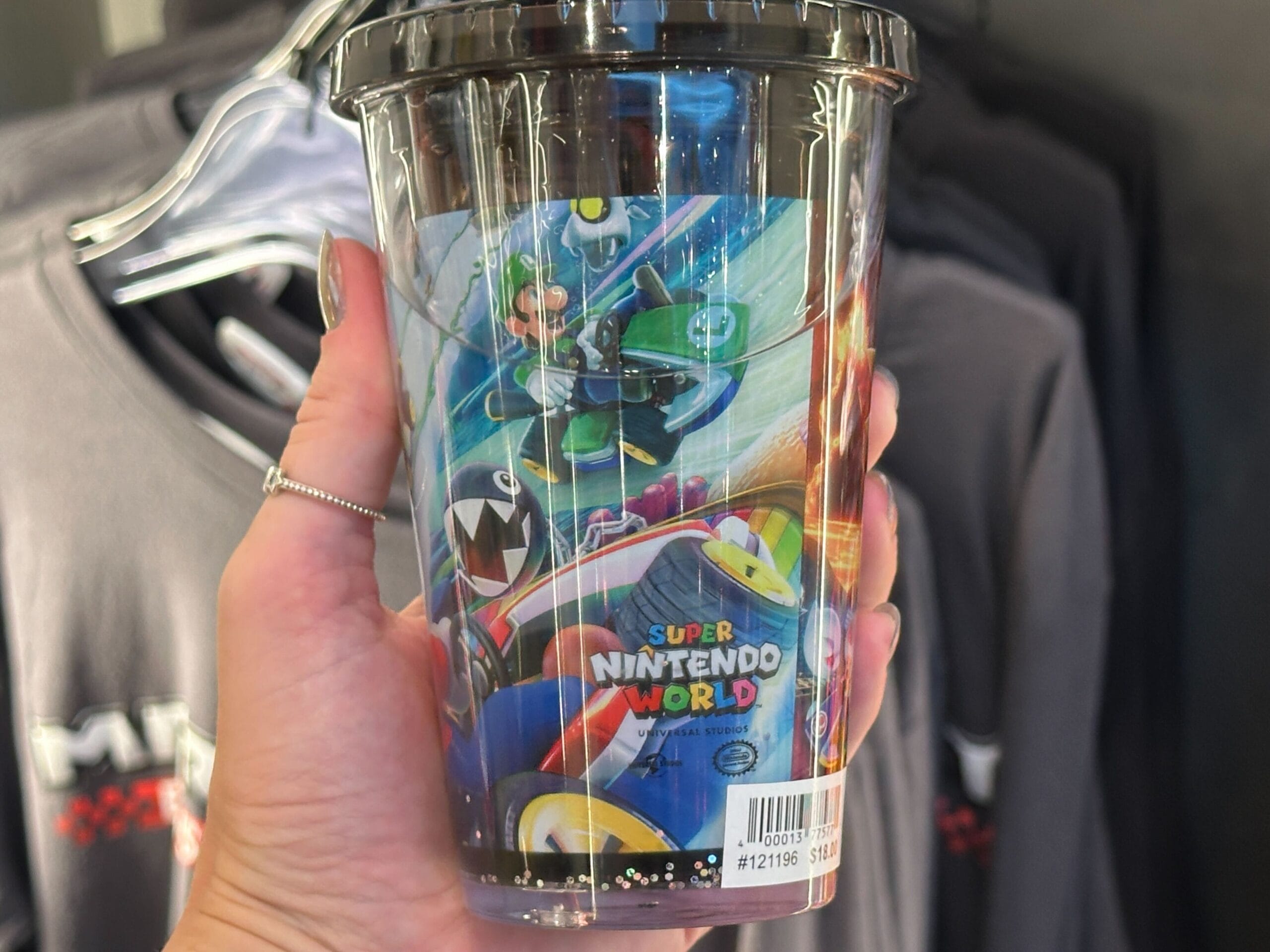 Hand holding a Super Nintendo World cup featuring Mario Kart characters, with Mario Motors merchandise shirts in the background.