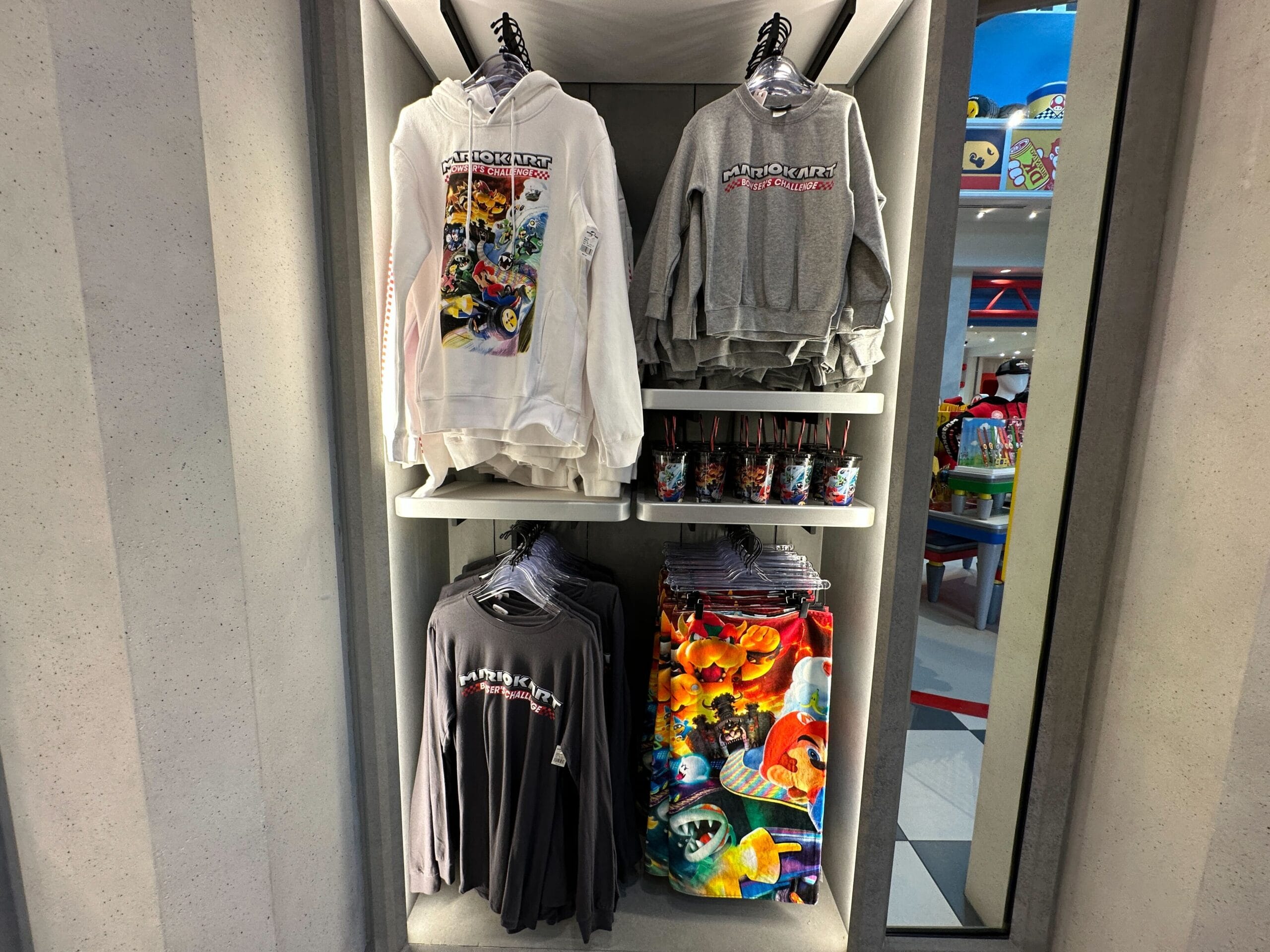 Super Nintendo World display with hoodies, shirts, cups, and Mario Motors merchandise featuring colorful Mario characters.