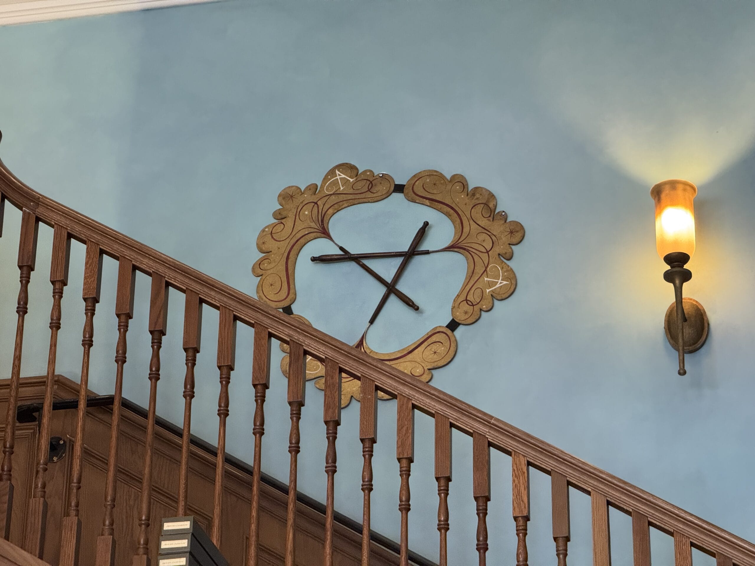 Gold decorative frame and Cosme Acajor baguettes magique hang on a blue wall above a wooden staircase, next to a lit sconce.