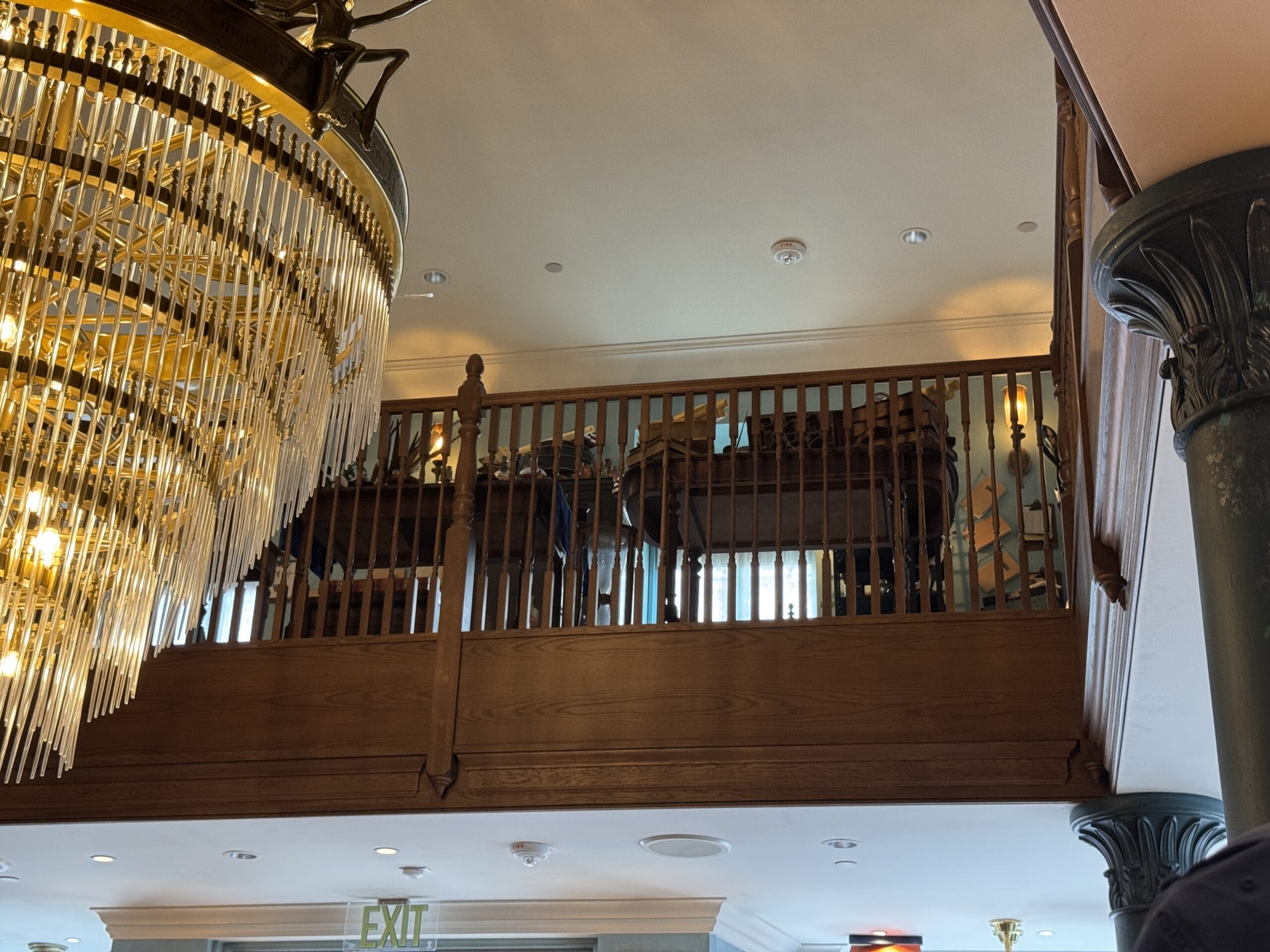 Large chandelier hangs near a wooden balcony with ship models and Cosme Acajor Baguettes Magique nautical decor.