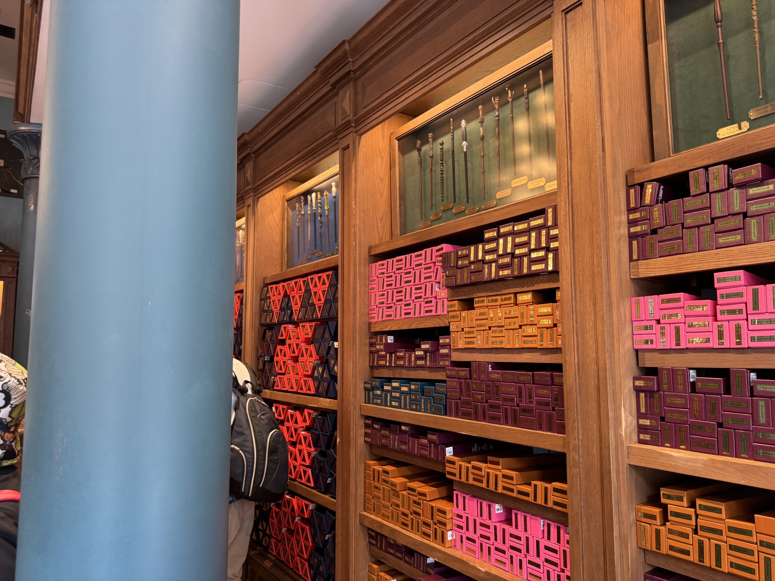 Shelves filled with Cosme Acajor Baguettes Magique; colorful wand boxes and wands line the walls, blue pillar in foreground.