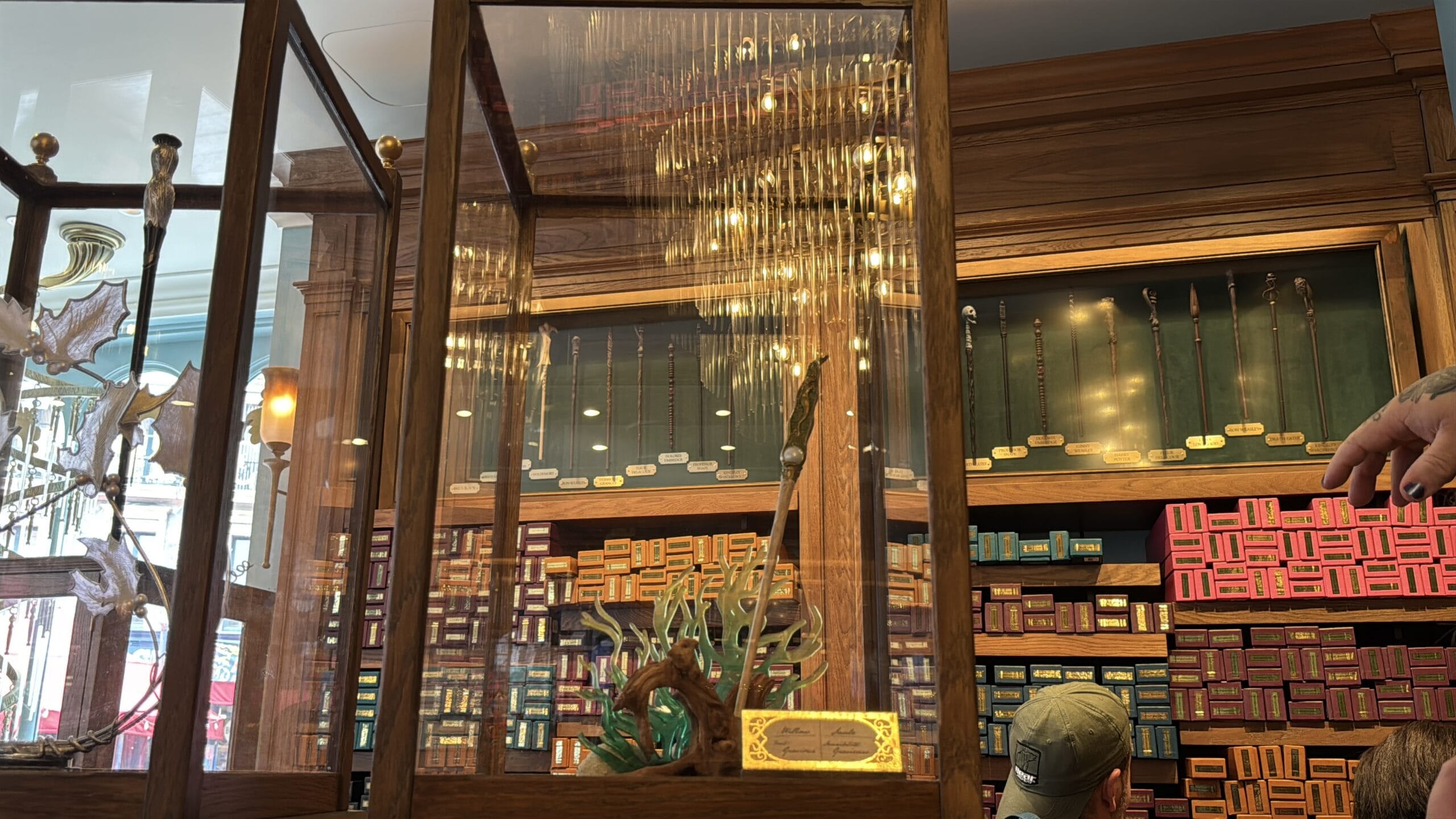 Cosme Acajor Baguettes Magique wands displayed in a glass case with shelves of colorful wand boxes behind.