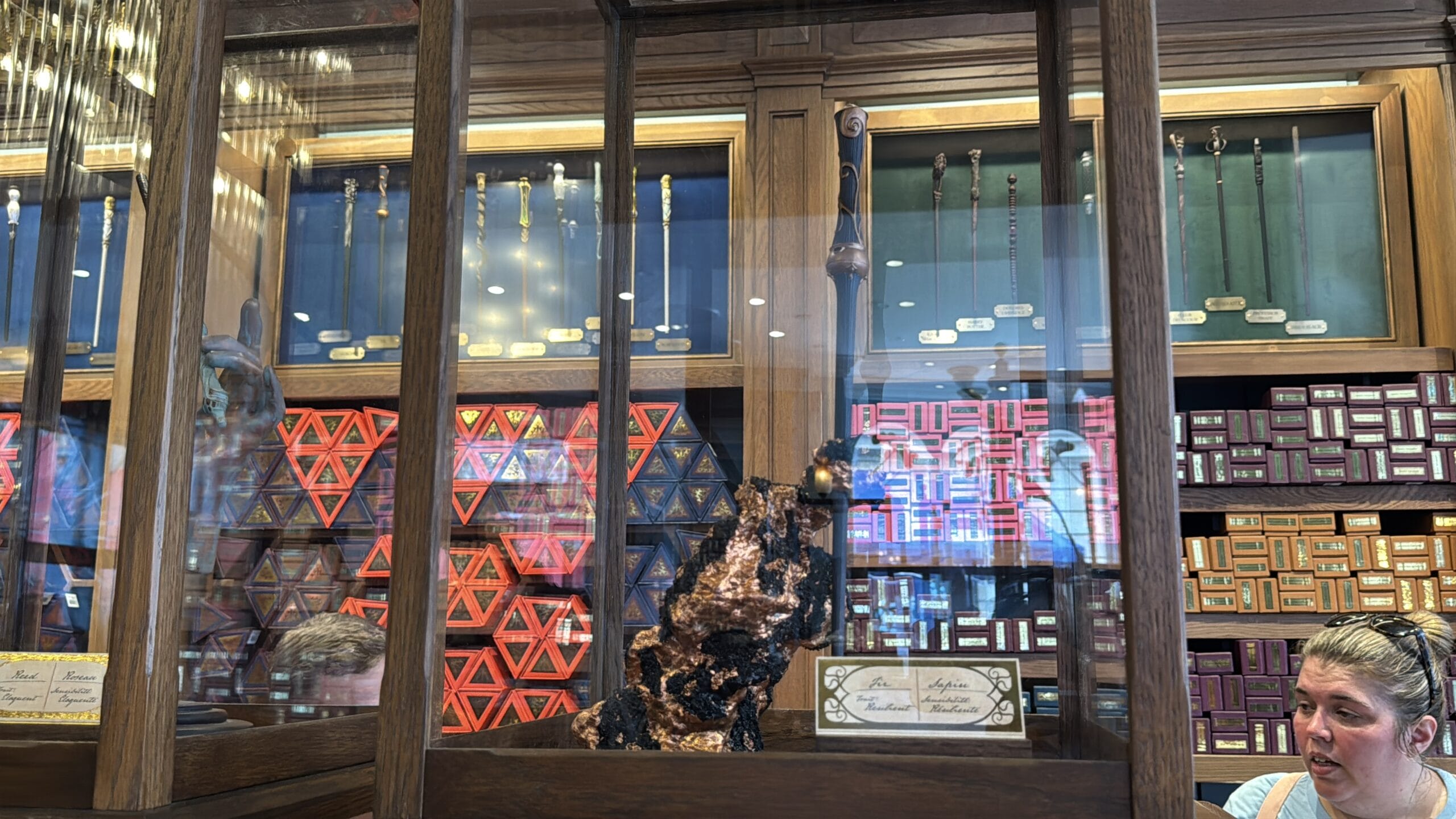 A display case at Cosme Acajor holds Baguettes Magique, with a large dark object and wand boxes nearby. Two people look on.