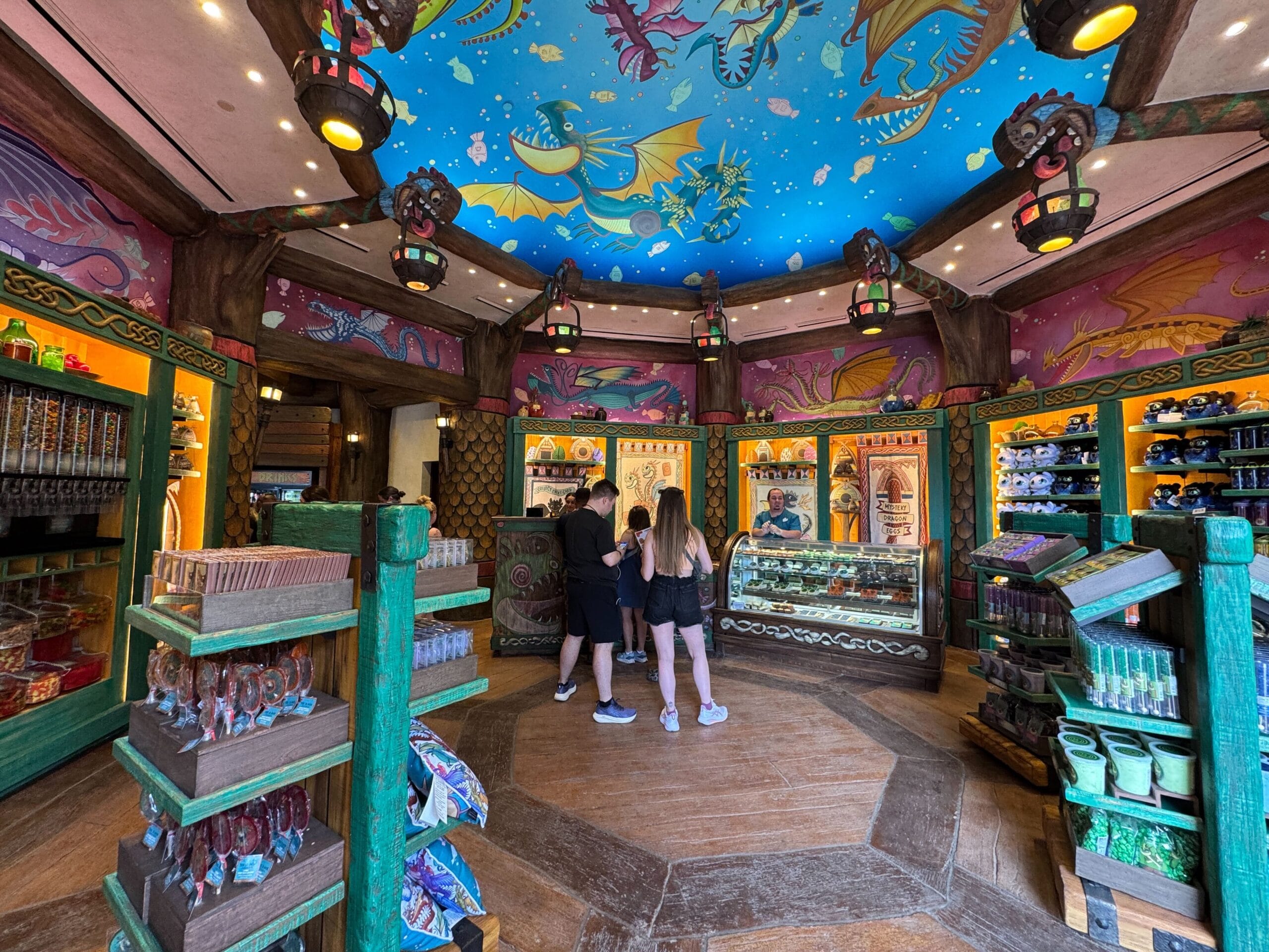 Colorful candy shop with fantasy-themed decor; three people explore treats beneath a How to Treat Your Dragon ceiling.