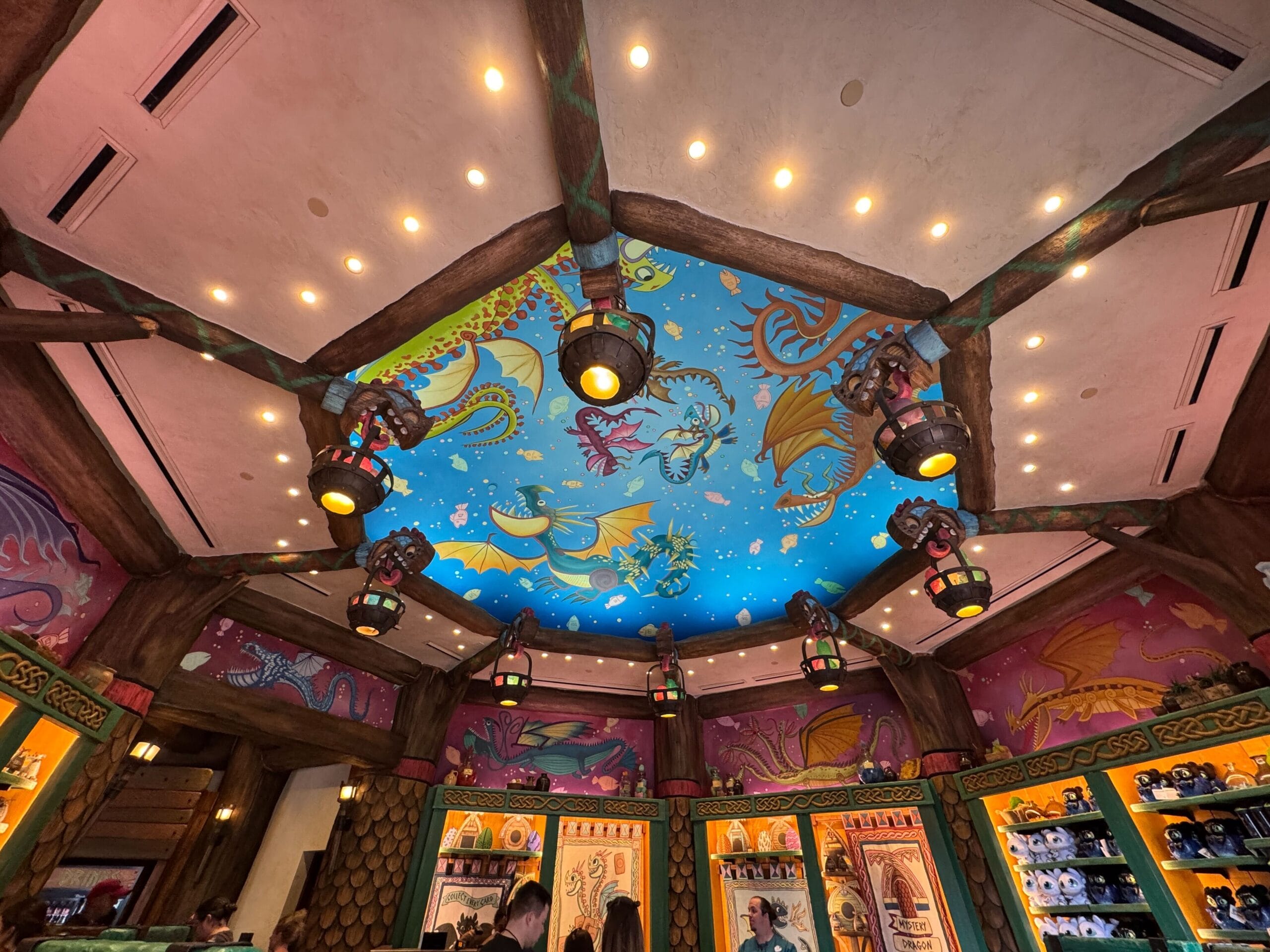 Colorful How to Treat Your Dragon mural on a domed ceiling with lanterns above a busy souvenir shop.