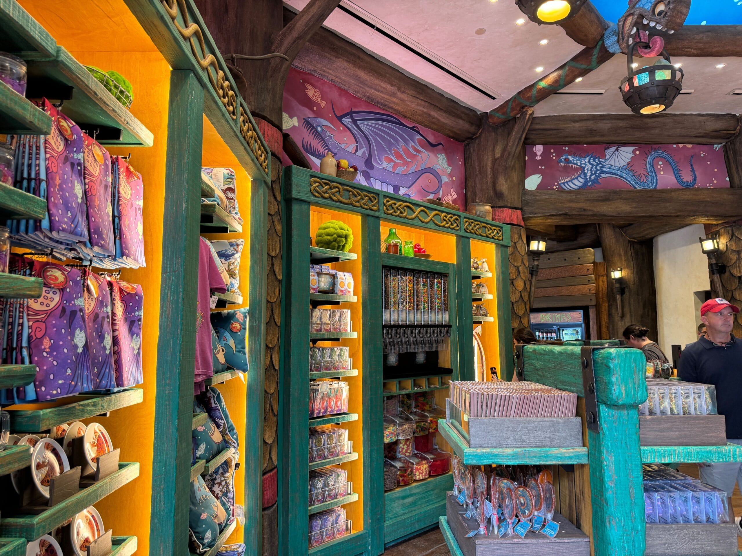 Colorful gift shop with How to Treat Your Dragon decor, shelves of toys, mugs, souvenirs, and two people in the background.