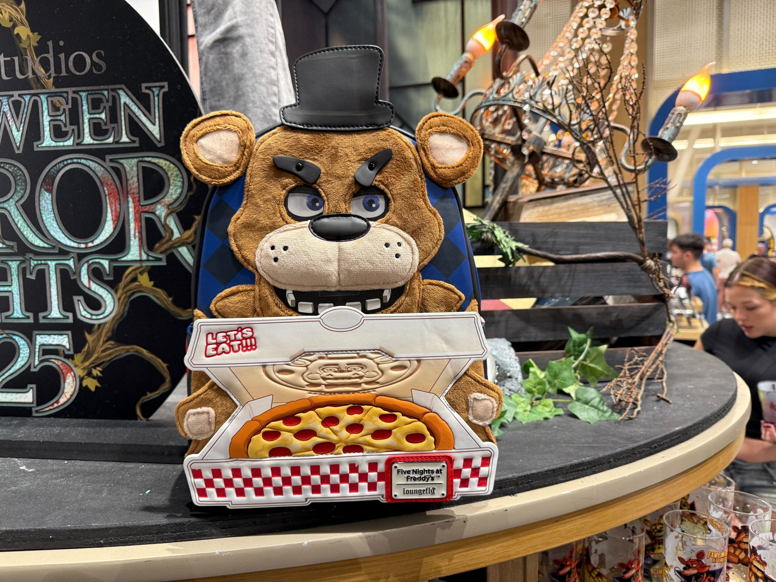 A plush bear Loungefly purse with a top hat holds a pizza box; FNAF Halloween Horror Nights 2025 decor is in the background.