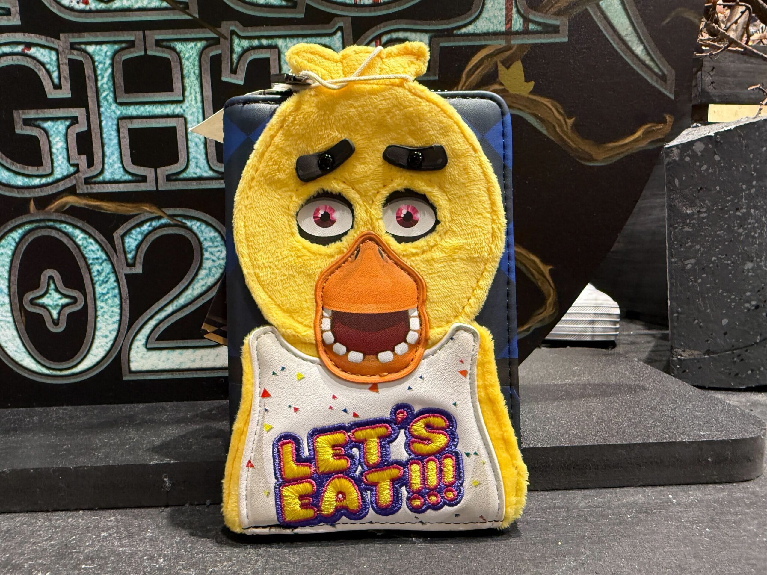 A Loungefly plush pouch shaped like Chica from FNAF with "LET'S EAT!!!" on its bib, displayed on a table.