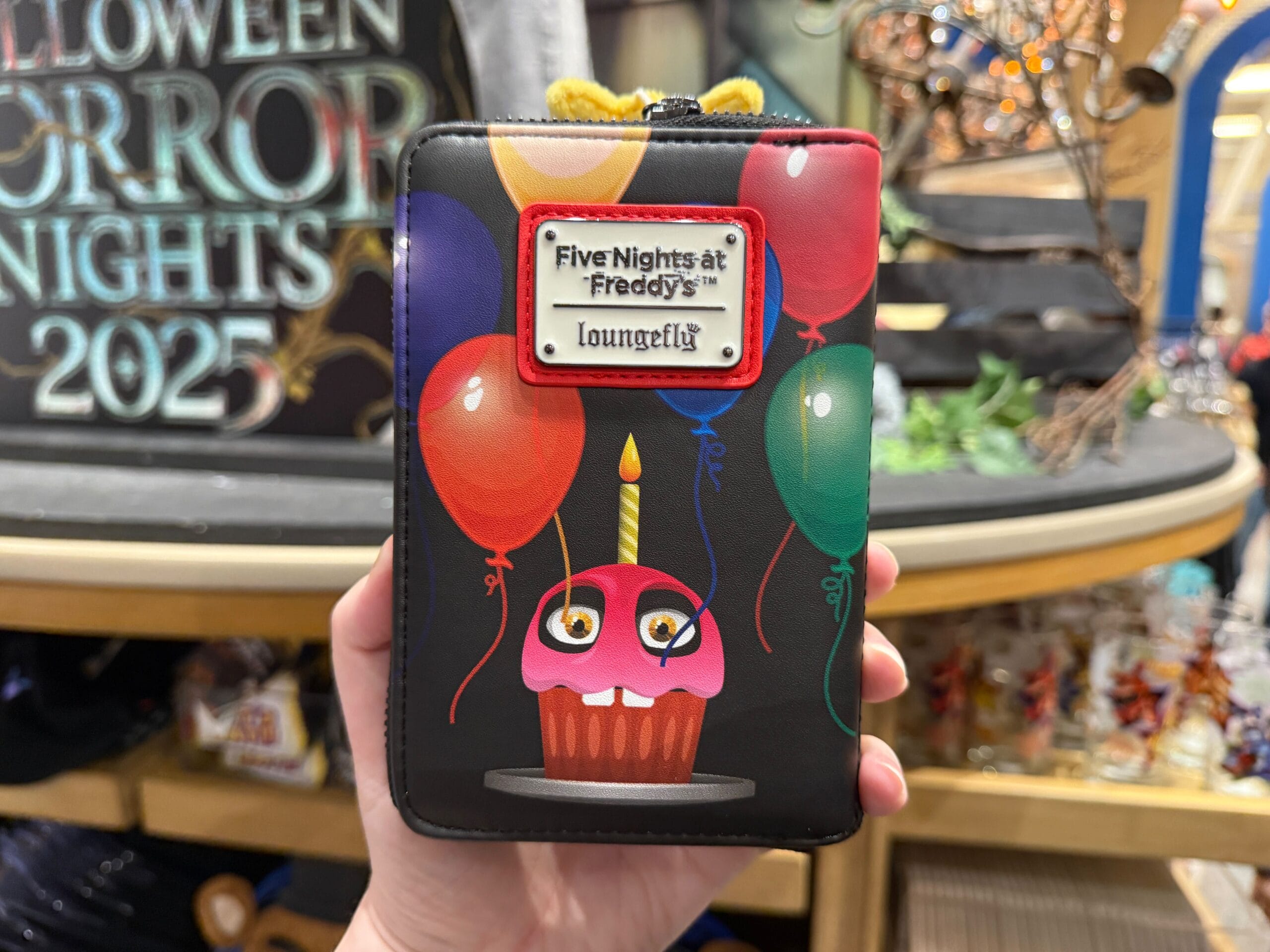 A hand holds an FNAF Loungefly wallet with a cupcake and colorful balloons design.