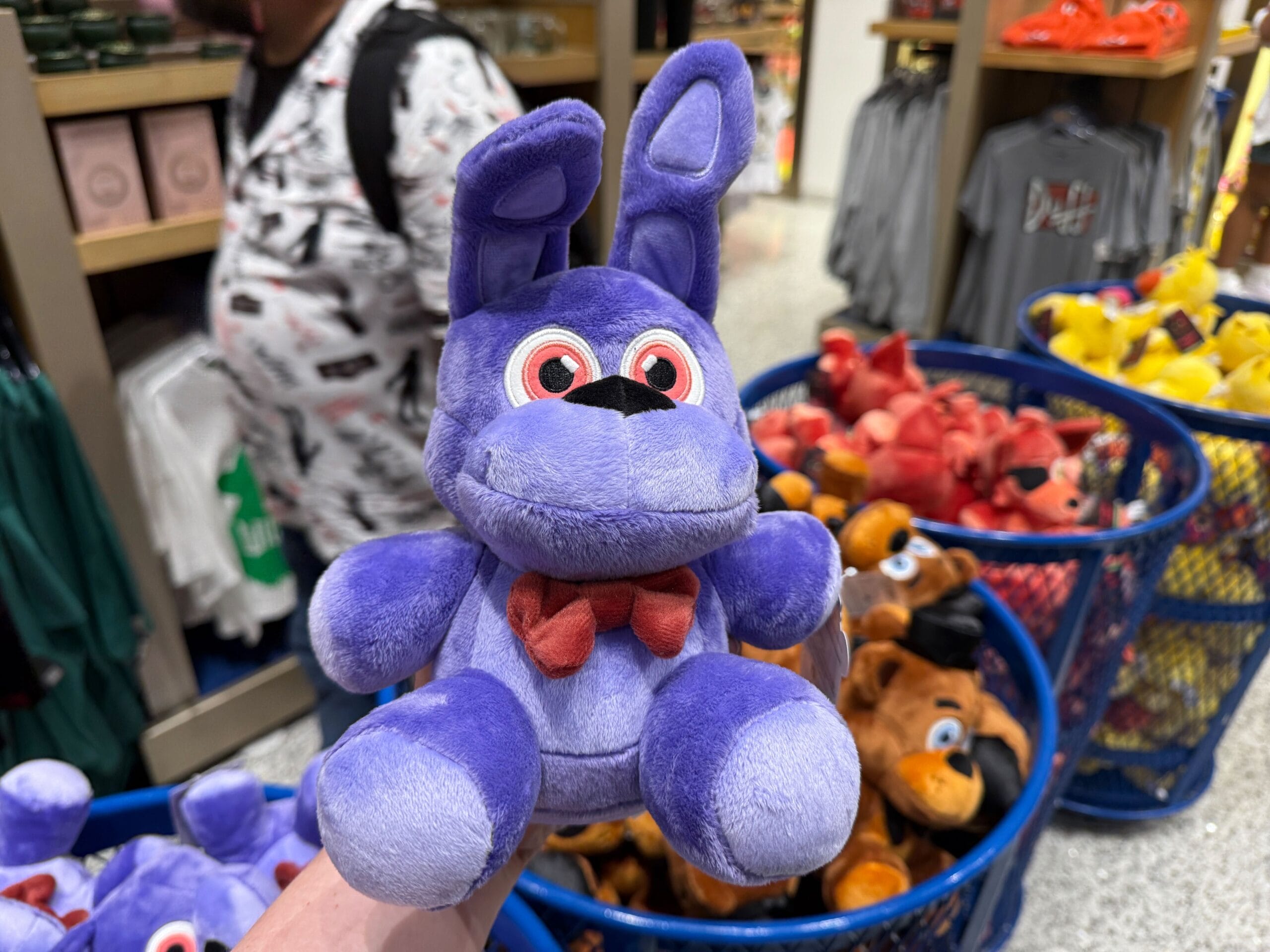 A hand holds a small purple plush bunny with a red bowtie, perfect for any FNAF or Loungefly collector, in a plush-filled store.