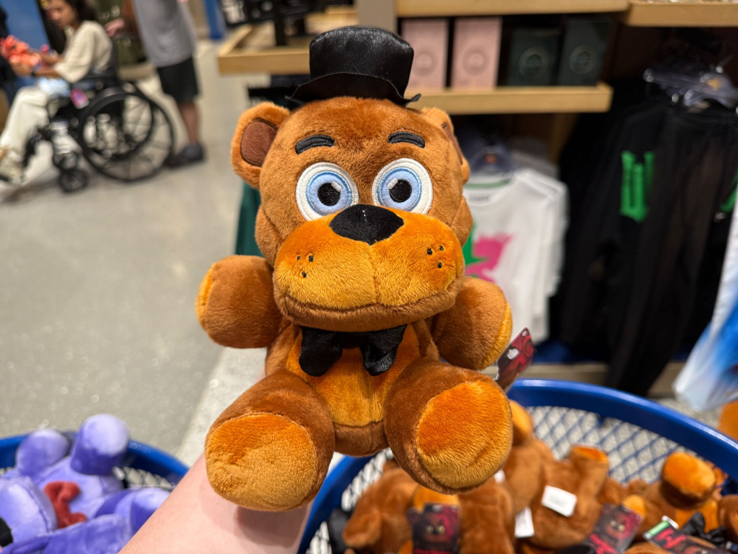 A hand holds a Loungefly FNAF plush bear toy with blue eyes, black bow tie, and top hat in a store.