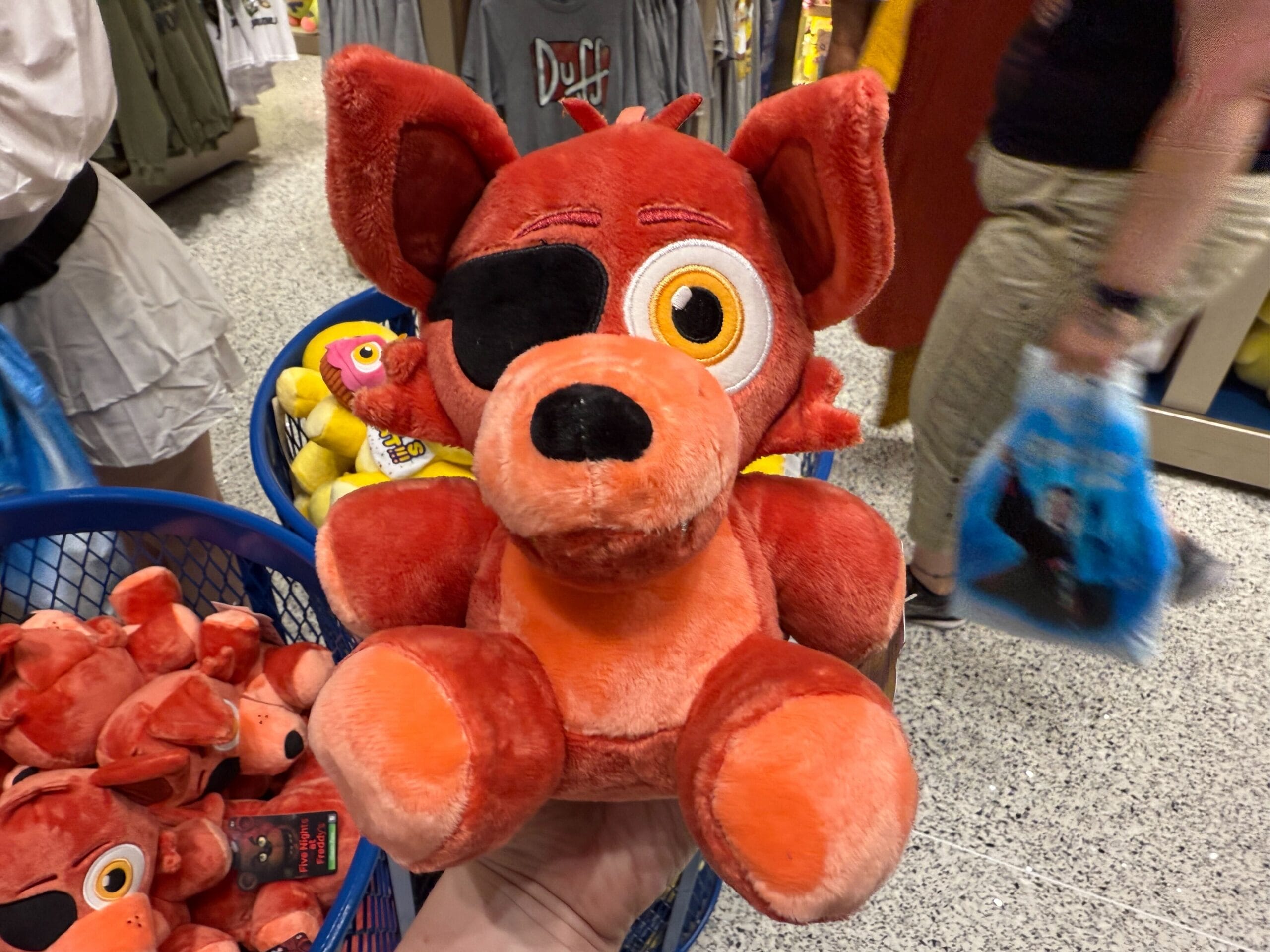A hand holds a red plush FNAF fox with an eye patch in a store, surrounded by Loungefly and other plush toys in baskets.