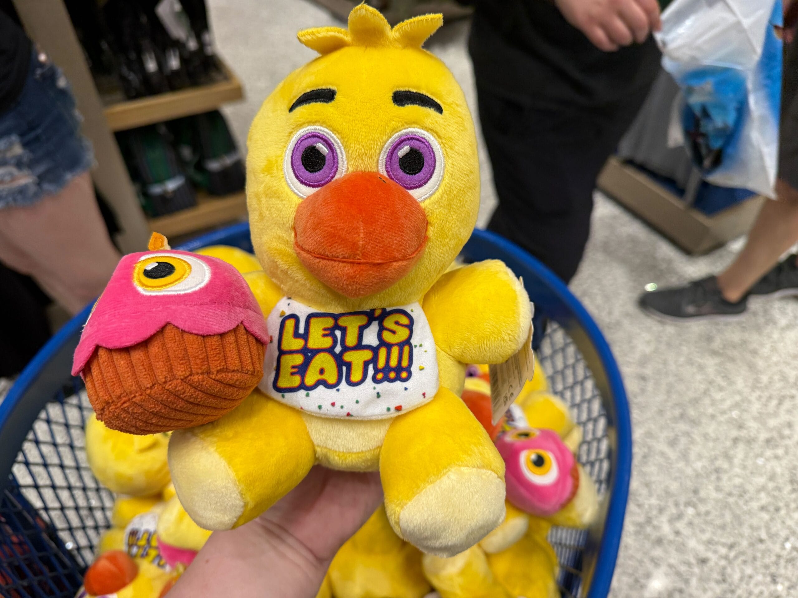 A hand holds a yellow FNAF "Let's Eat!!!" plush duck with a cupcake, surrounded by toys in a store basket.