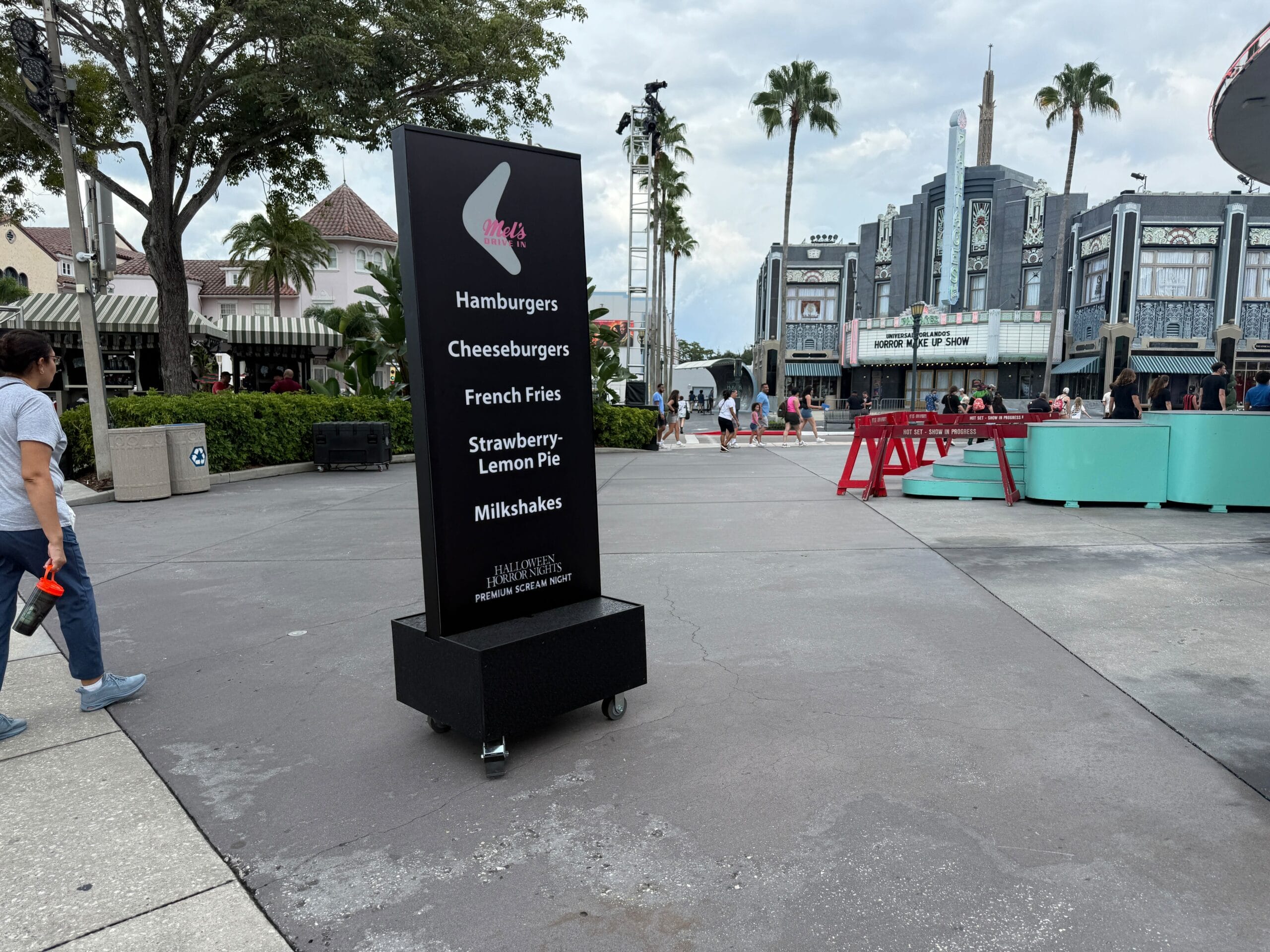 A black menu sign at an outdoor plaza for Halloween Horror Nights lists burgers, fries, pie, and milkshakes.