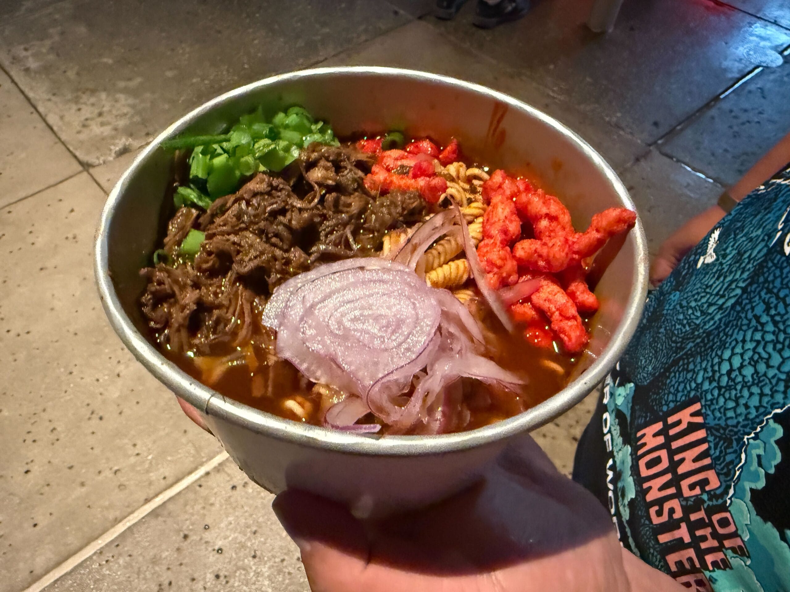 A hand holds a bowl of ramen topped with beef and onions at Halloween Horror Nights: perfect for your review.