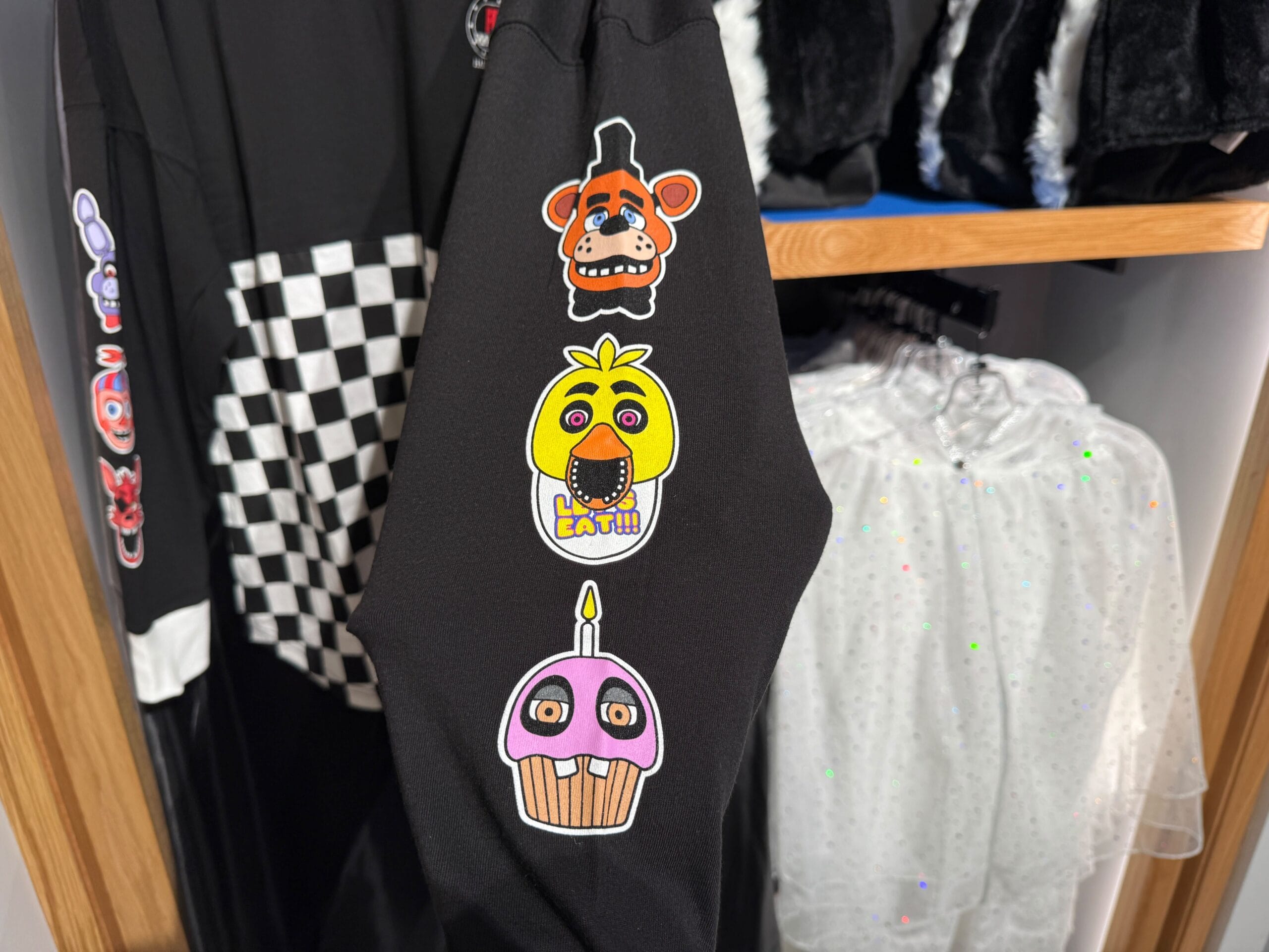 Black sleeve with cartoon patches of a bear, chicken, and cupcake, hanging on a rack with other clothes.