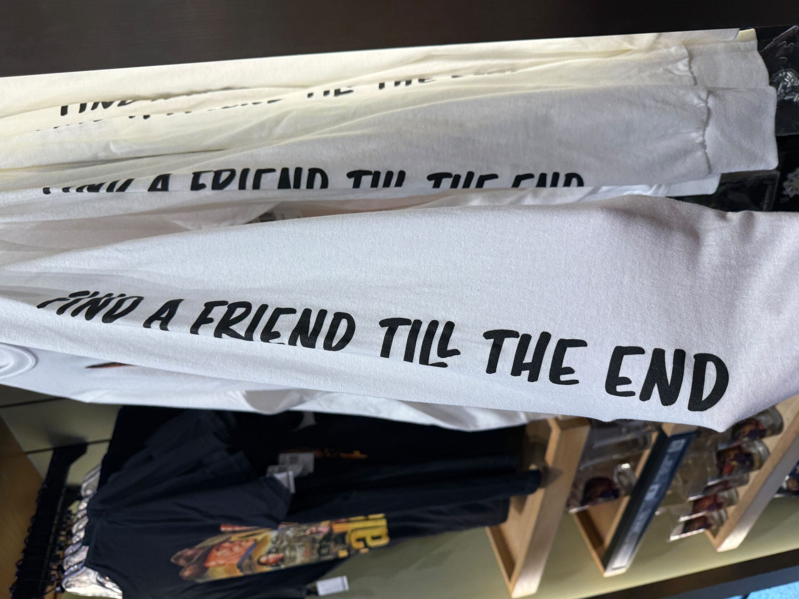 White long-sleeve shirts on a rack, printed with the text “FIND A FRIEND TILL THE END” in bold black letters.