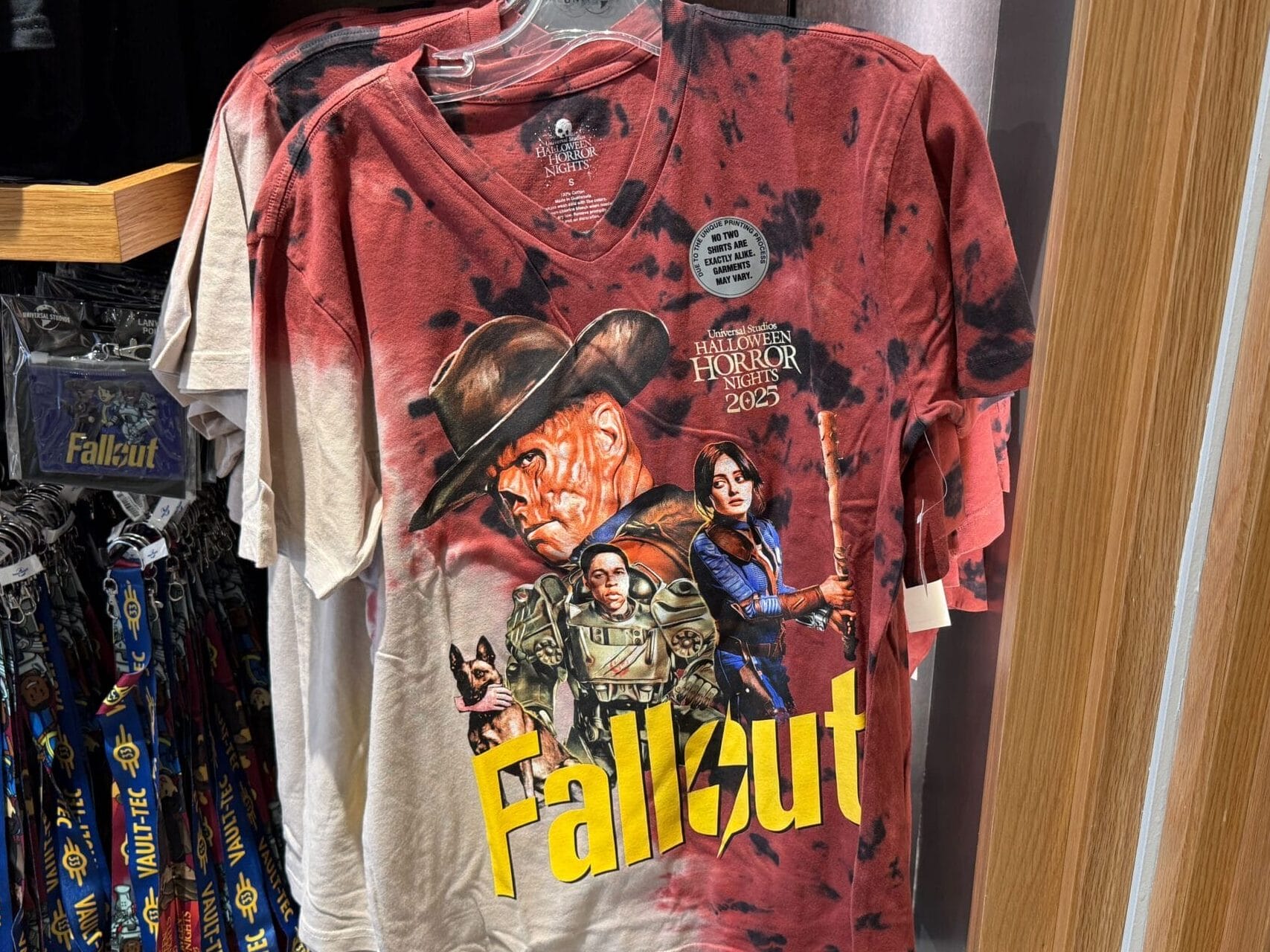 A red and white Fallout T-shirt with game characters hangs on display in a store next to Fallout-branded lanyards.