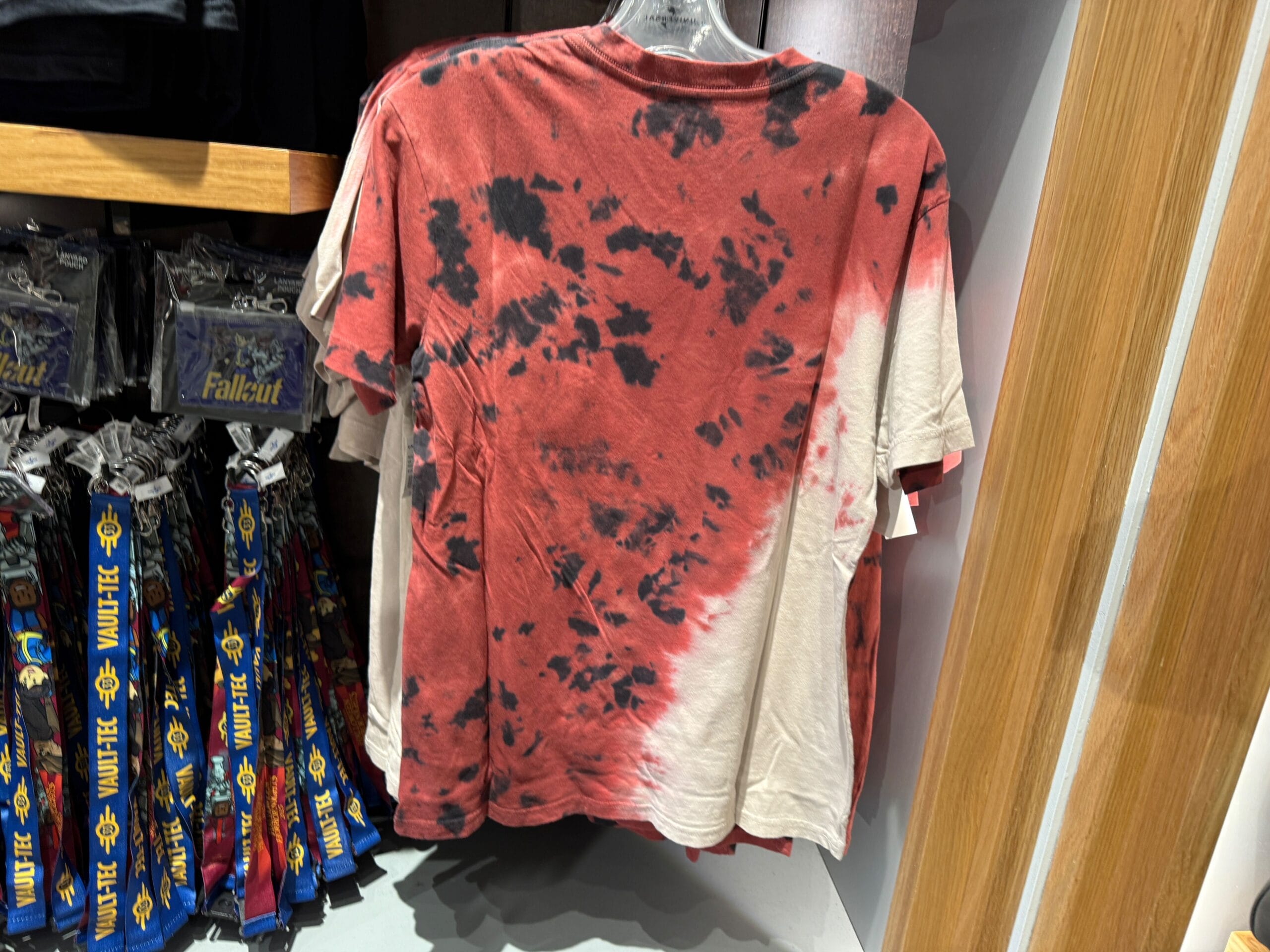 Red and black tie-dye T-shirt hanging on a rack, next to Fallout-themed lanyards in a retail store.