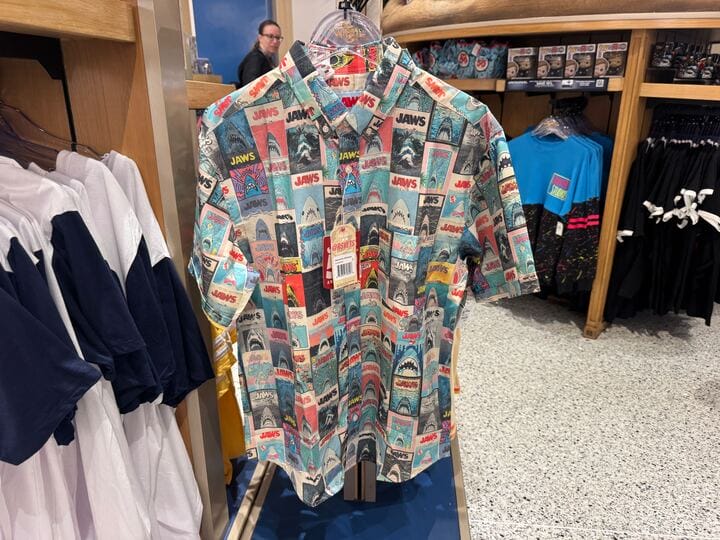 Short-sleeve button-up shirt covered in colorful vintage Jaws movie poster designs, hanging in a store display.