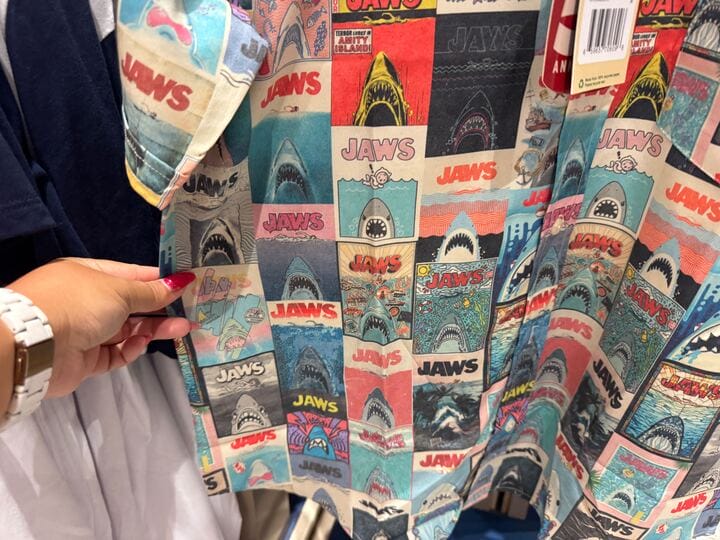 A hand holds a shirt printed with colorful Jaws movie posters featuring sharks and the word "JAWS.