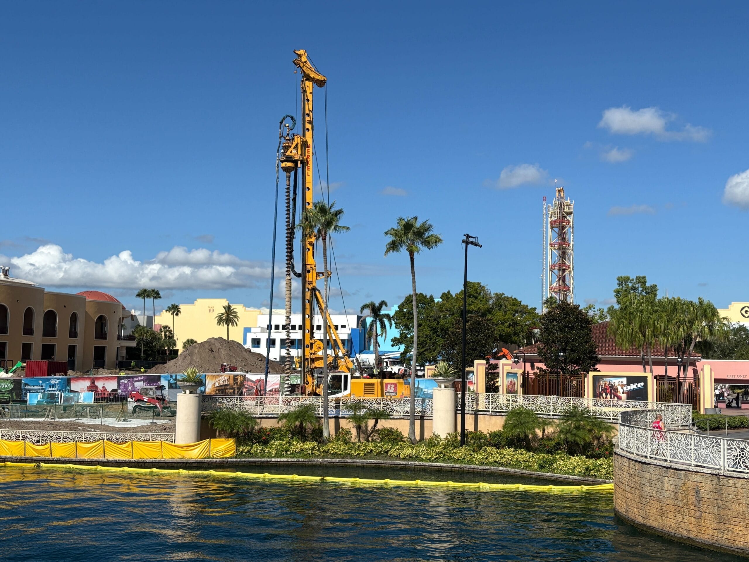 A large drilling machine at a construction site near water, with palm trees and Rip Ride Rockit in the background.