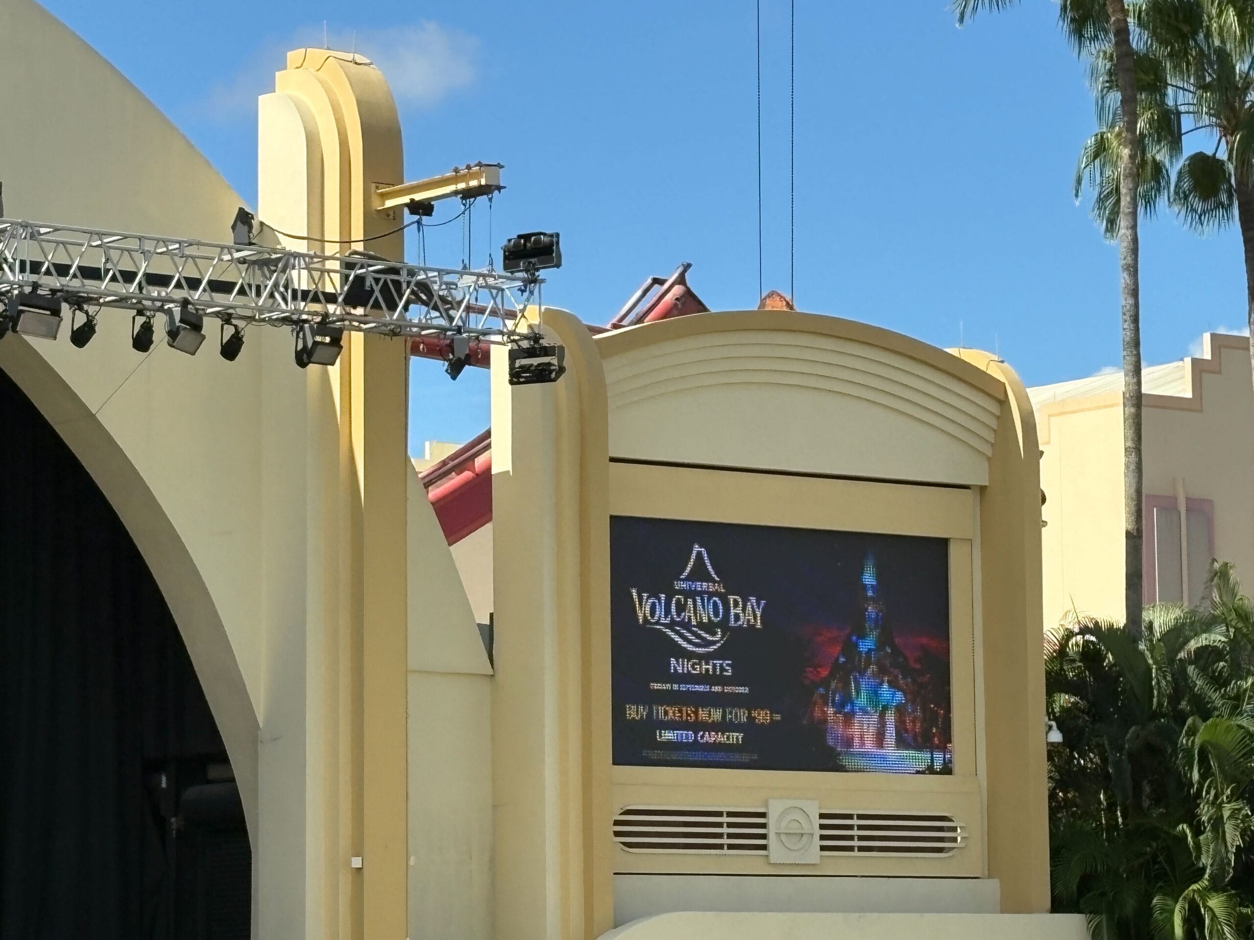 Large outdoor stage at Universal Studios with a digital sign for Volcano Bay Nights under palm trees and blue sky.