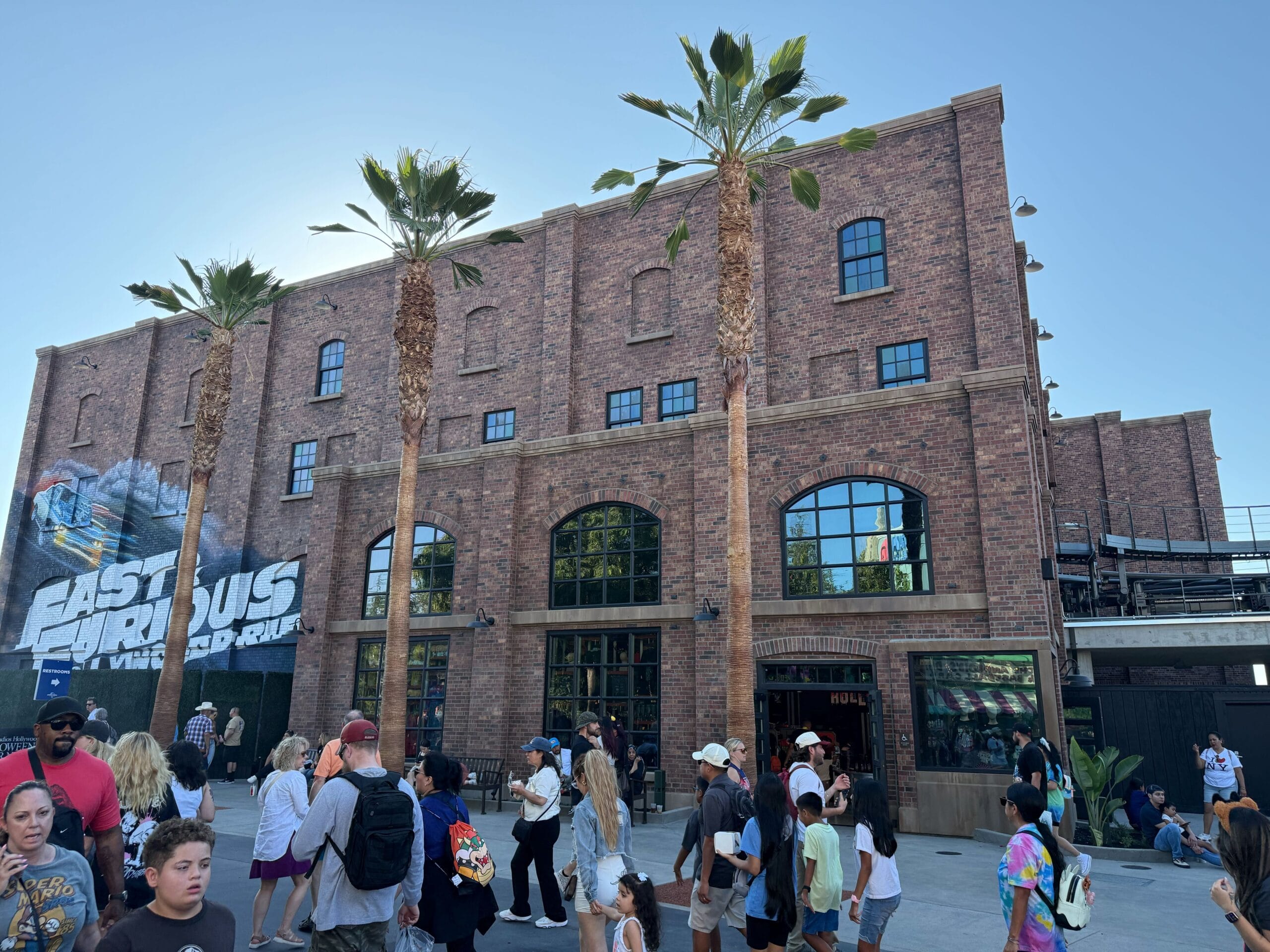 Groups of people walk by a brick building with palm trees and a Fast & Furious Store at Universal Studios Hollywood.