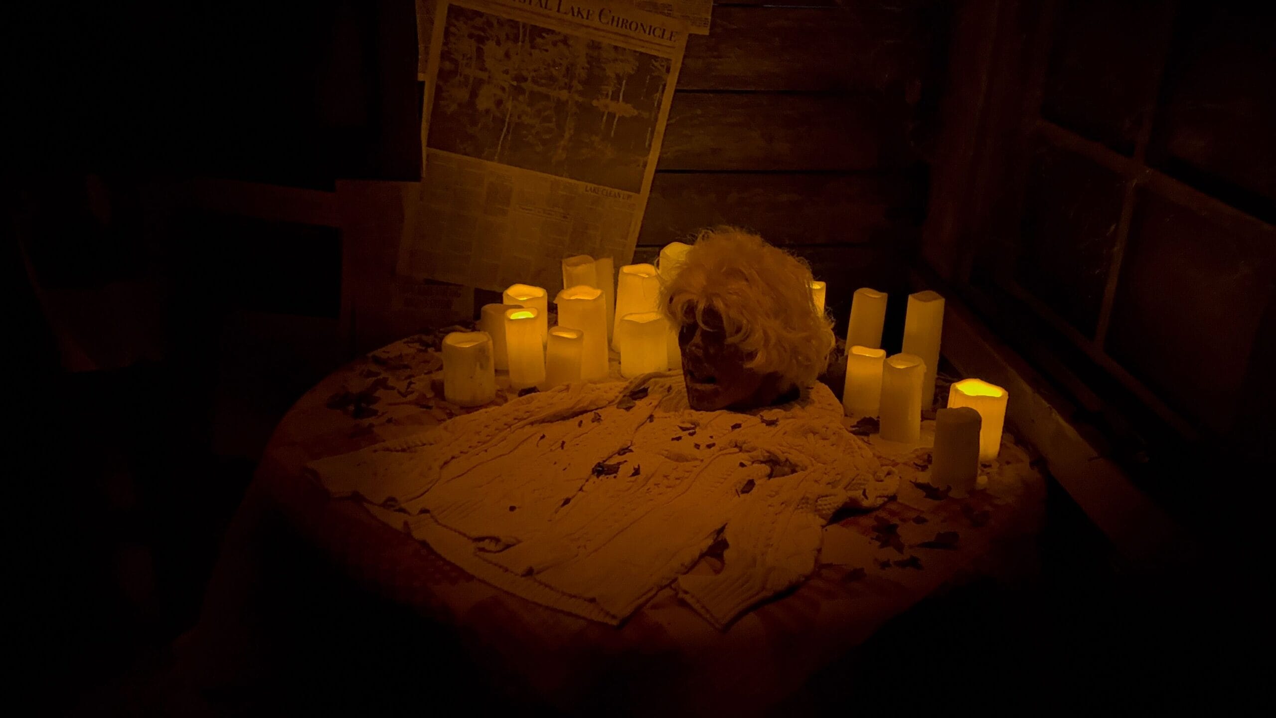 A decayed head on a sweater surrounded by lit candles on a table in a dim, rustic room with a newspaper on the wall.