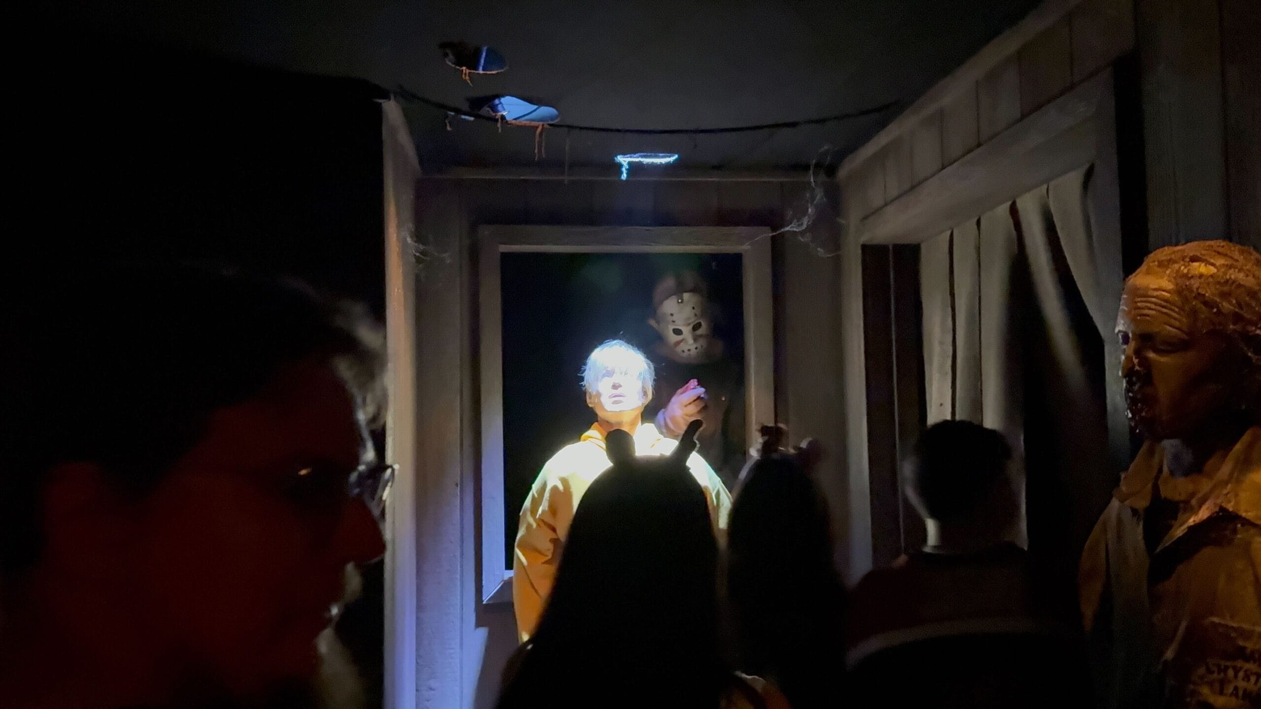 People walk through a dark room featuring a glowing figure in a yellow raincoat and a mask hanging above their head.
