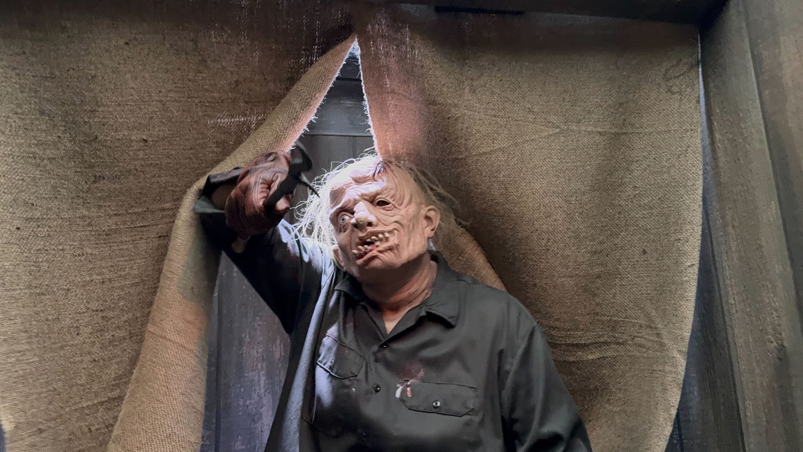 A person in a scary monster mask and dark clothes stands in front of torn burlap curtains.