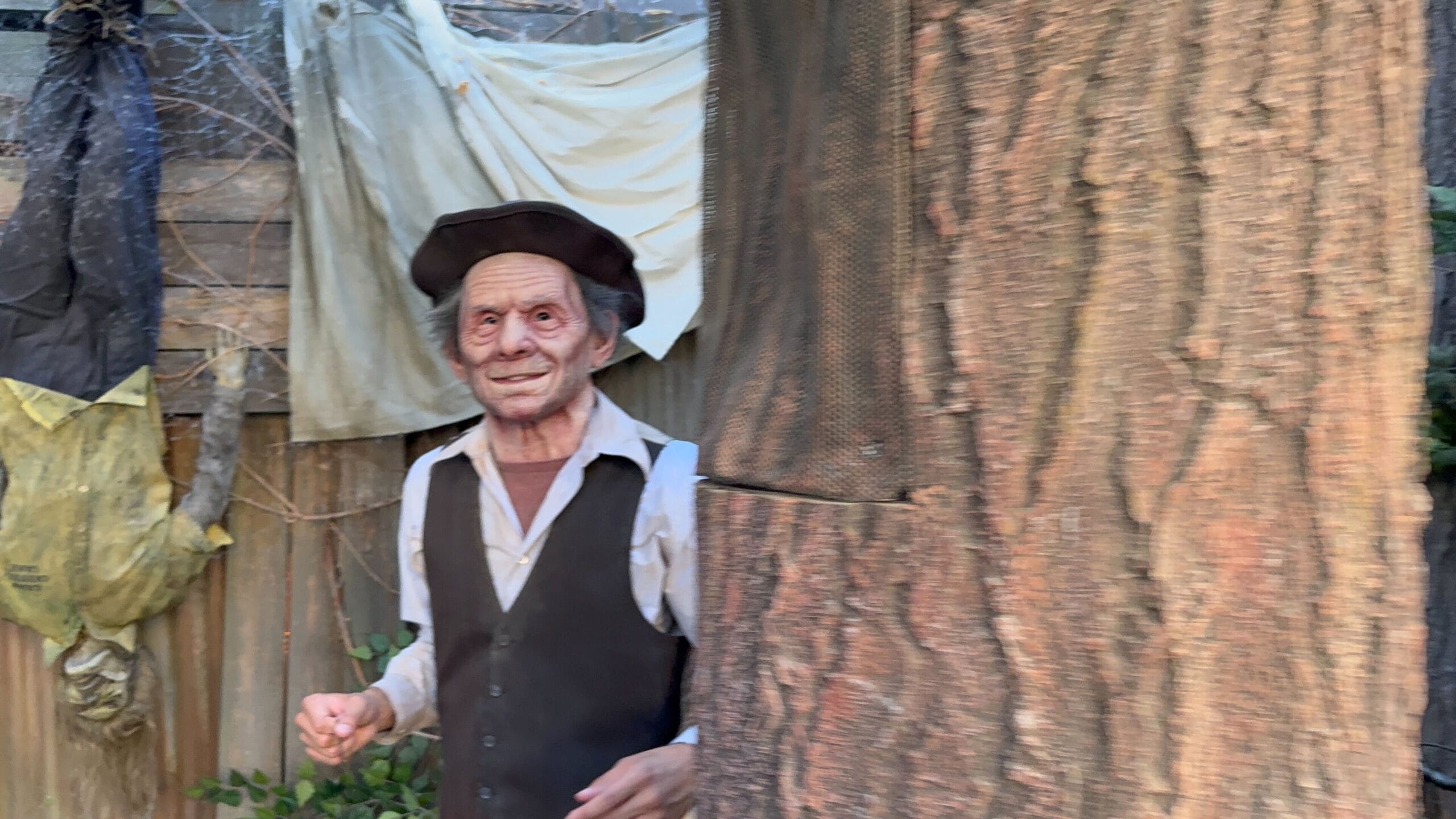 A creepy, life-sized mannequin in old-fashioned clothes and hat stands by a large tree in a yard with Halloween decorations.
